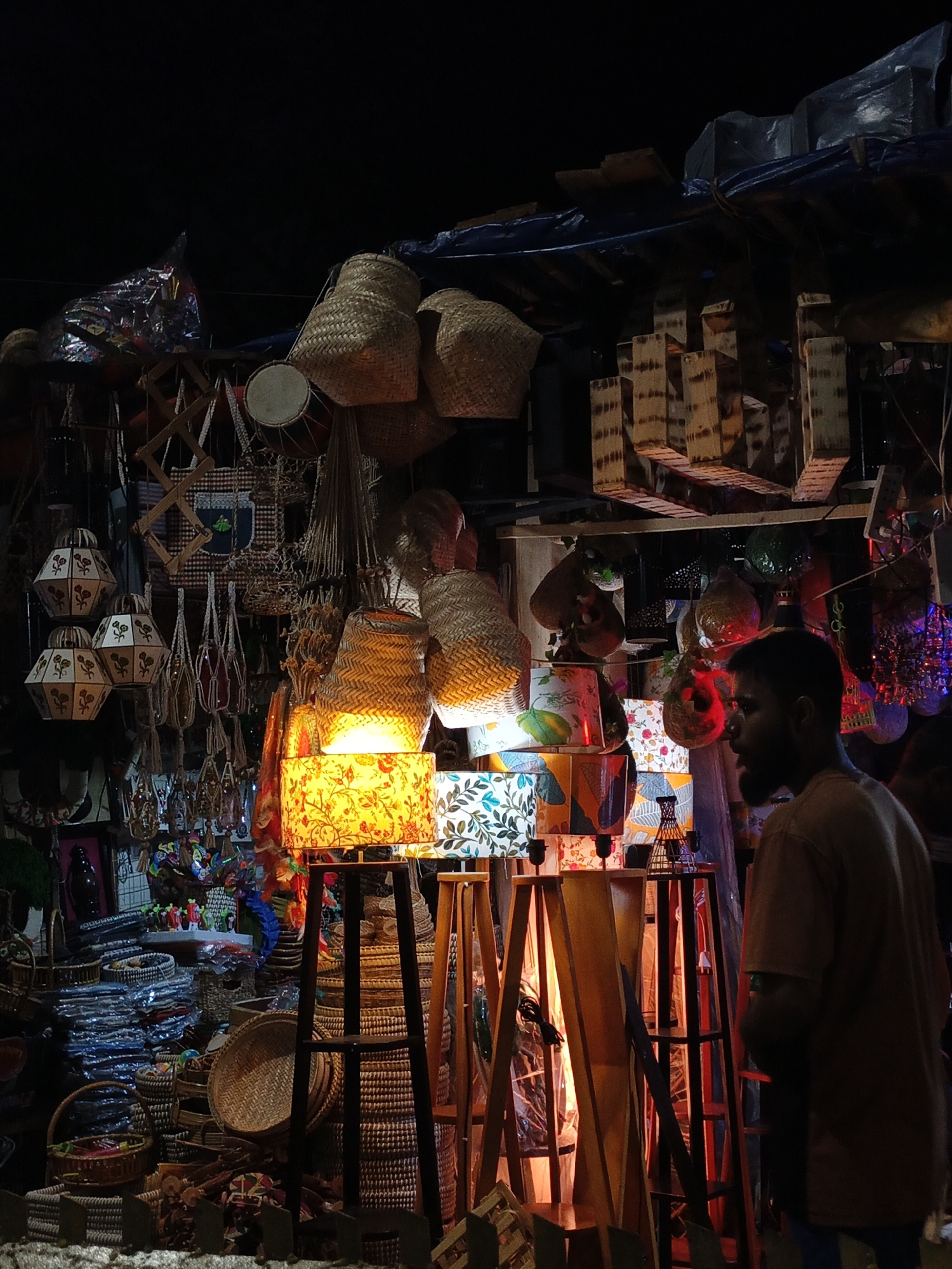 A nighttime scene in Dhaka, Bangladesh, shows an outdoor market stall packed with various handmade crafts. The stall is dimly lit with colorful lamps and hanging lights, illuminating woven baskets, lampshades with floral patterns, and wooden items. A man stands to the right of the frame, his face partially obscured, wearing a brown shirt. Stacks of baskets are visible on the left side of the stall. The stall is covered by a tarp-like structure, with various items hanging overhead.