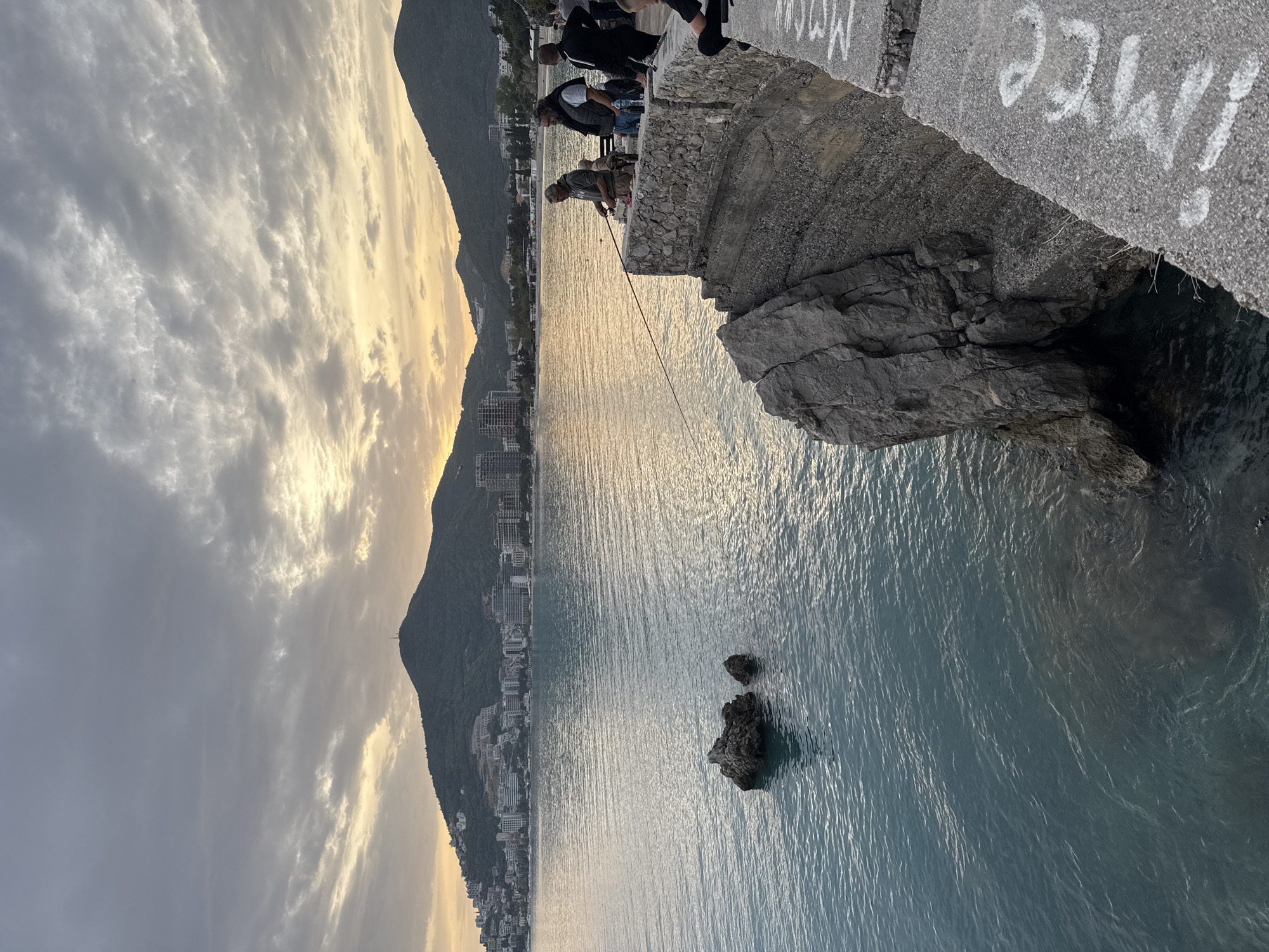 The image, taken with a vertical orientation, captures a picturesque coastal scene, likely depicting Budva, Montenegro, at either sunrise or sunset. The sky is partly cloudy, showcasing a warm golden glow near the horizon that gently reflects on the calm, rippling surface of the sea.
Along the distant coastline, a lush mountain range rises, its lower slopes densely packed with numerous buildings that form a vibrant coastal town. In the foreground, a rugged concrete and rock pier juts out into the water. Several individuals are gathered on this structure. A man in a light-colored shirt is prominently featured, actively engaged in fishing with a rod extending over the water. Behind him, another person in a dark jacket stands, seemingly observing. Further back, a small group of people are also visible, enjoying the tranquil setting.
Scattered in the calm waters near the pier are a few dark, craggy rocks. On the concrete surface of the pier, white graffiti-like text is visible, appearing to spell out "MMCE!" or similar characters. The scene conveys a sense of peaceful activity, with people enjoying the evening or morning by the sea.