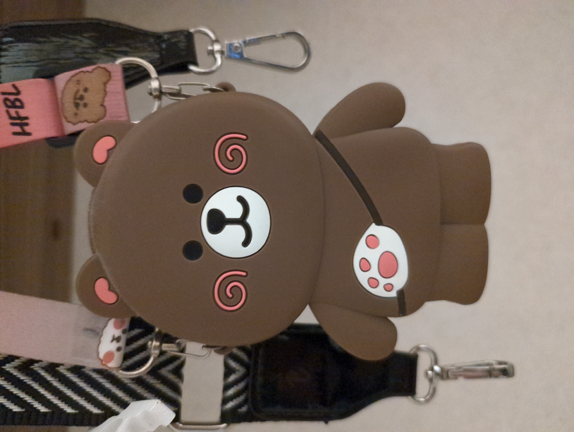 A close-up shot depicts a brown cartoon bear keychain. The bear has a large, round head with pink swirls on its cheeks, a white and black nose, and pink ears. Its body is connected with a black strap, and it features a white spot with pink paw prints. The keychain is attached to several straps, including a pink one with a brown bear print and the letters "HFBL," and a black and white zigzag patterned strap. A silver clip is also attached to the keychain. The background is a neutral, light color. The location is Unknown, Unknown.