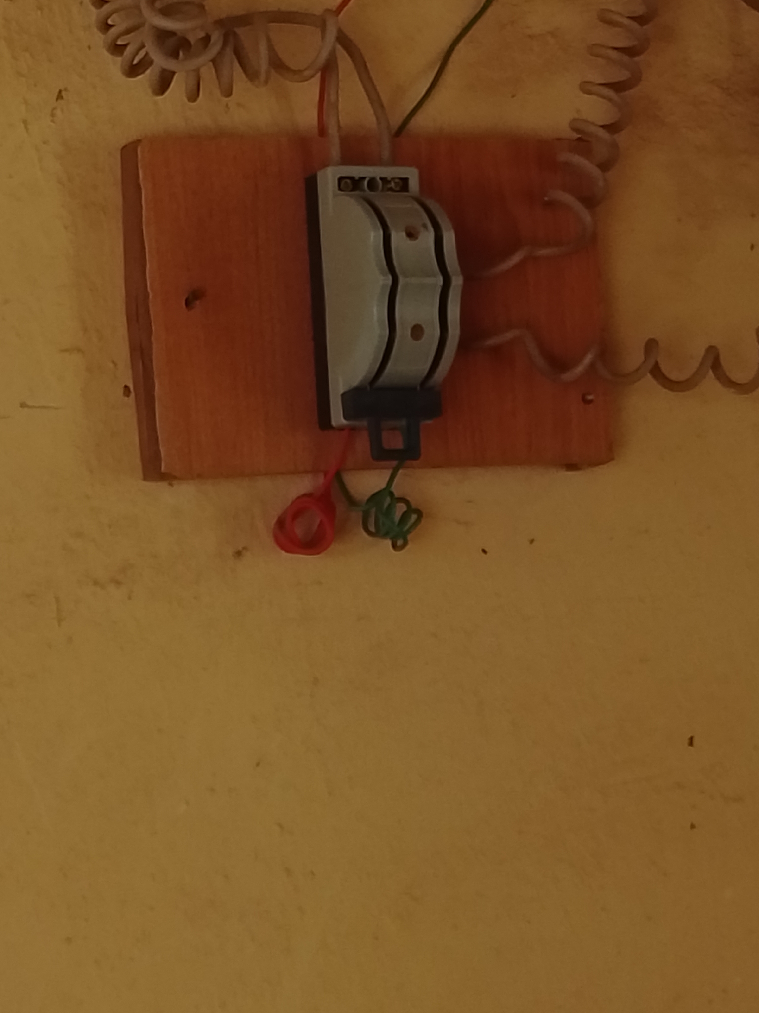 A close-up, eye-level shot shows a wall-mounted electrical switch and wiring in Jalingo, Nigeria. The switch, a light grey, double-pole unit with a black lever, is attached to a rectangular, light brown wooden board. Red and green insulated wires connect to the switch. Above the switch, a coiled grey wire is visible, attached to the wall. To the right of the switch, a similar coiled grey wire descends. The background wall is a textured, yellowish-brown color, with some subtle discoloration and debris. The lighting is dim, casting soft shadows.