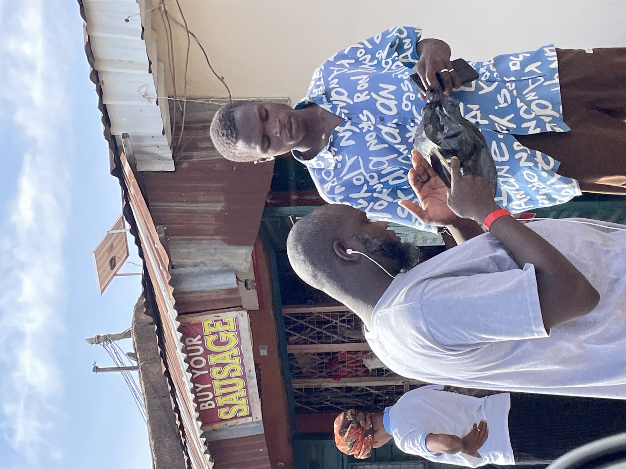 Two men are examining a black plastic bag. The man on the right wears a blue patterned shirt and brown pants. The man in front wears a white t-shirt, has a trimmed beard, and an earphone in his left ear. Behind them is a building with a sign that says "BUY YOUR SAUSAGE," along with a building with a satellite dish. The image appears to be taken in Jos, Nigeria.