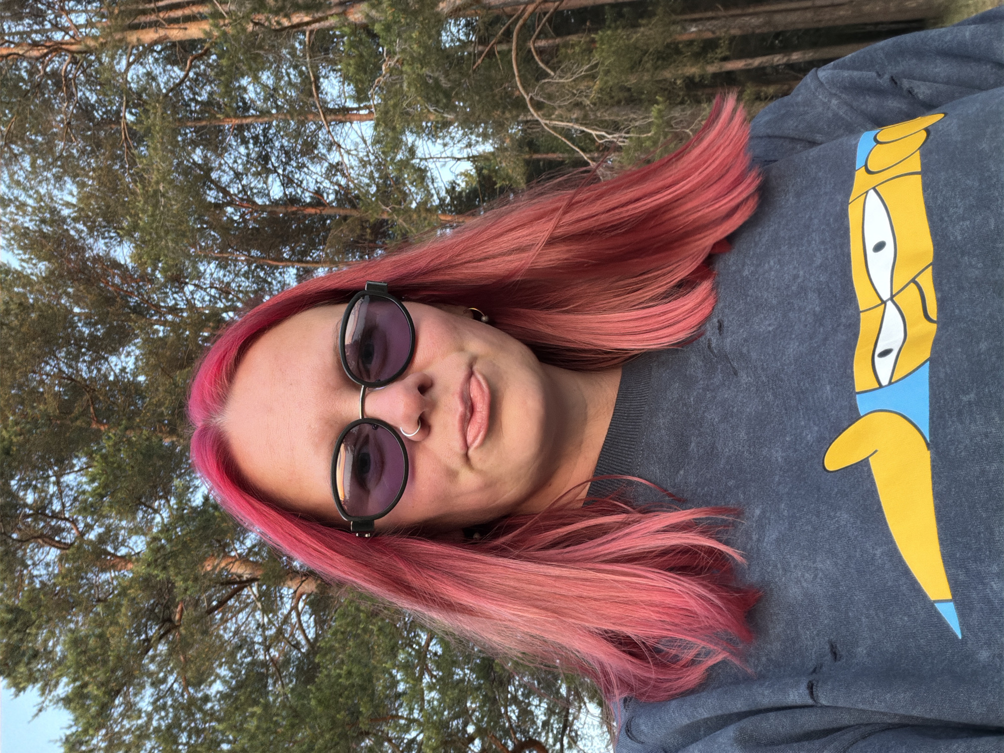 A young woman with vibrant pink hair and sunglasses poses for a selfie outdoors. She is wearing a dark grey sweatshirt with a cartoon graphic. The background consists of tall pine trees and a clear sky, suggesting a natural, possibly forested, environment. The lighting indicates it is late afternoon or early evening.