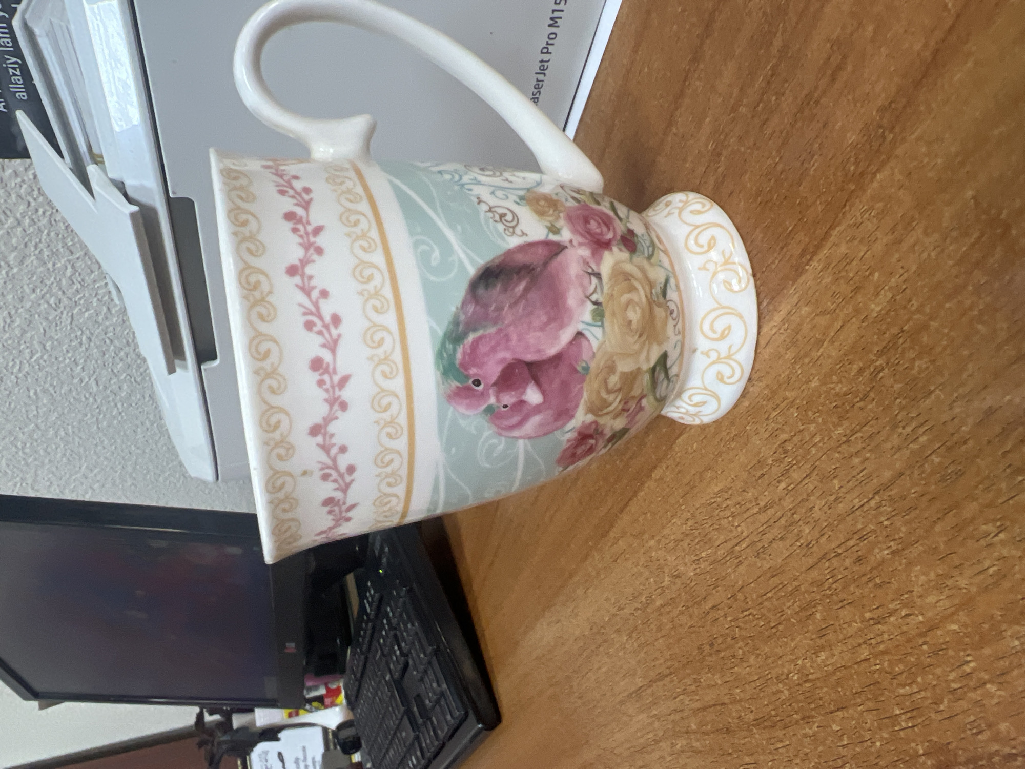 A close-up, eye-level shot shows a decorative mug with a floral and bird design on a wooden desk. The mug is white with pink and gold accents, featuring two pink birds perched among roses. To the left of the mug, a black computer keyboard is visible. In the background, a computer monitor and a portion of a printer can be seen. The desk surface is made of wood with a visible grain pattern. The lighting is soft, casting no harsh shadows.