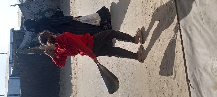 The image captures an outdoor scene in Khan Yunis, Palestinian Territory, under daylight conditions. Two individuals are depicted from behind, walking towards the right side of the frame. In the foreground, a person, appearing to be a child or young individual, wears a red hooded sweatshirt, dark gray or black sweatpants, and light beige open-toe slides. This individual holds a dark, elongated object, possibly a rolled blanket or bag, by its gathered end in their left hand, with their right arm slightly raised. To their right and slightly behind, an adult female wears a full black abaya that covers her head and body, holding a clear plastic bag containing light-colored items, possibly food, in her left hand. The ground surface consists of packed dirt or sand, with a lighter, possibly paved, horizontal strip in the lower portion. In the background, several makeshift structures are visible, constructed from dark tarpaulins, corrugated metal sheets, and wooden supports. One structure features a black fabric panel adorned with white text or designs. Elongated shadows of the two individuals are cast to their left on the ground, indicating a light source from the right.