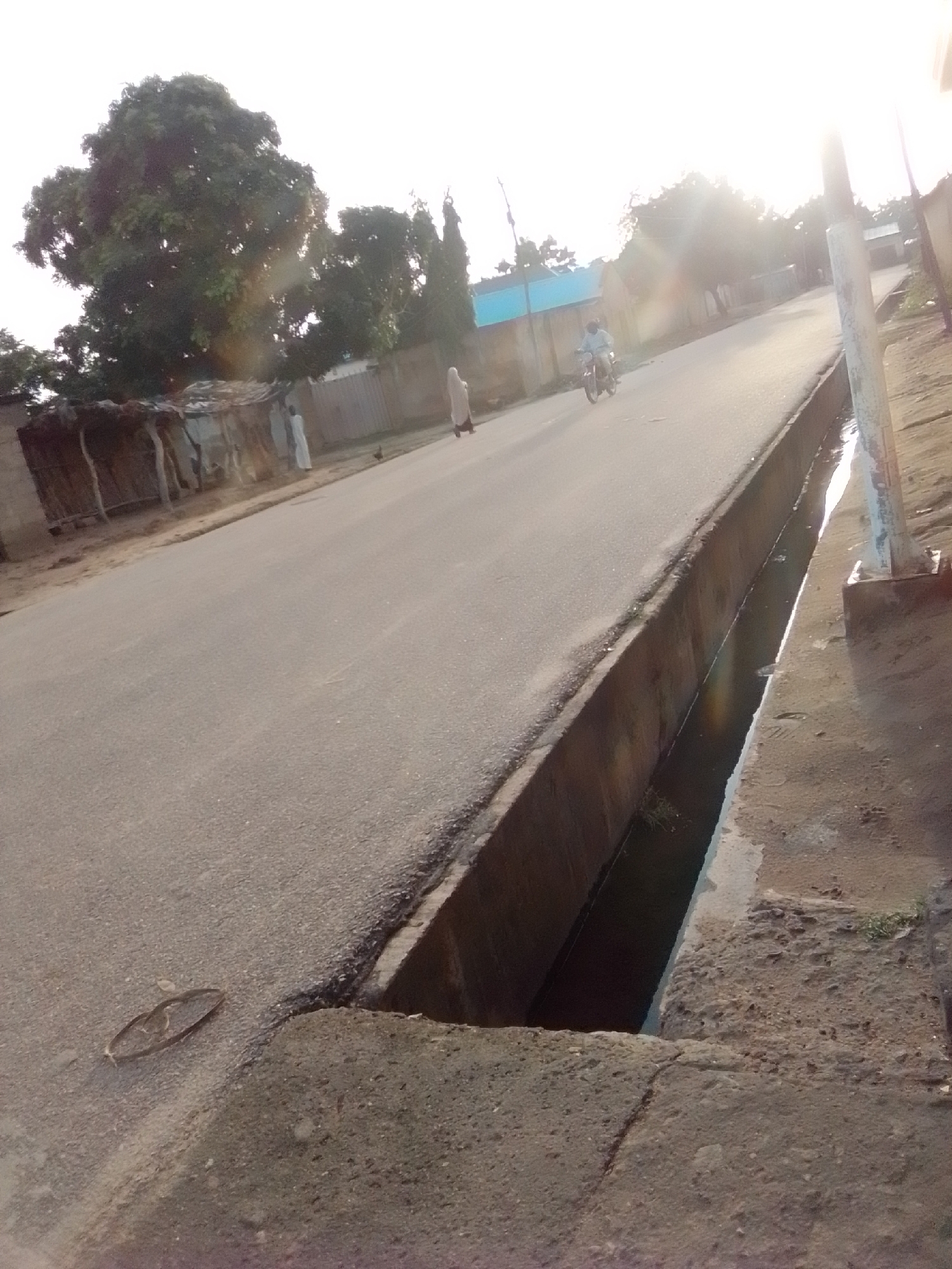 The image depicts a street scene in Gombe, Nigeria, characterized by a paved road with a concrete drainage channel running alongside it. The sun is low in the sky, casting long shadows and creating lens flare. On the left side of the road, there are structures with corrugated metal roofs and what appear to be mud walls, suggesting a residential or commercial area. A large, leafy tree dominates the left portion of the frame.
Several individuals are visible. A person in a light-colored garment is walking on the road. Another individual is riding a motorcycle. A person in a white robe is standing near the structures on the left. A chicken is also present near the structures.
The road surface shows signs of wear. A circular object, possibly a lid or cover, is lying on the asphalt near the drainage channel. The drainage channel itself contains some water and vegetation. The ground beside the drainage channel is rough and appears to be dirt and concrete. In the background, there are buildings and more vegetation, with a bright, washed-out sky. The overall impression is a typical street in a Nigerian city during the late afternoon or early morning.