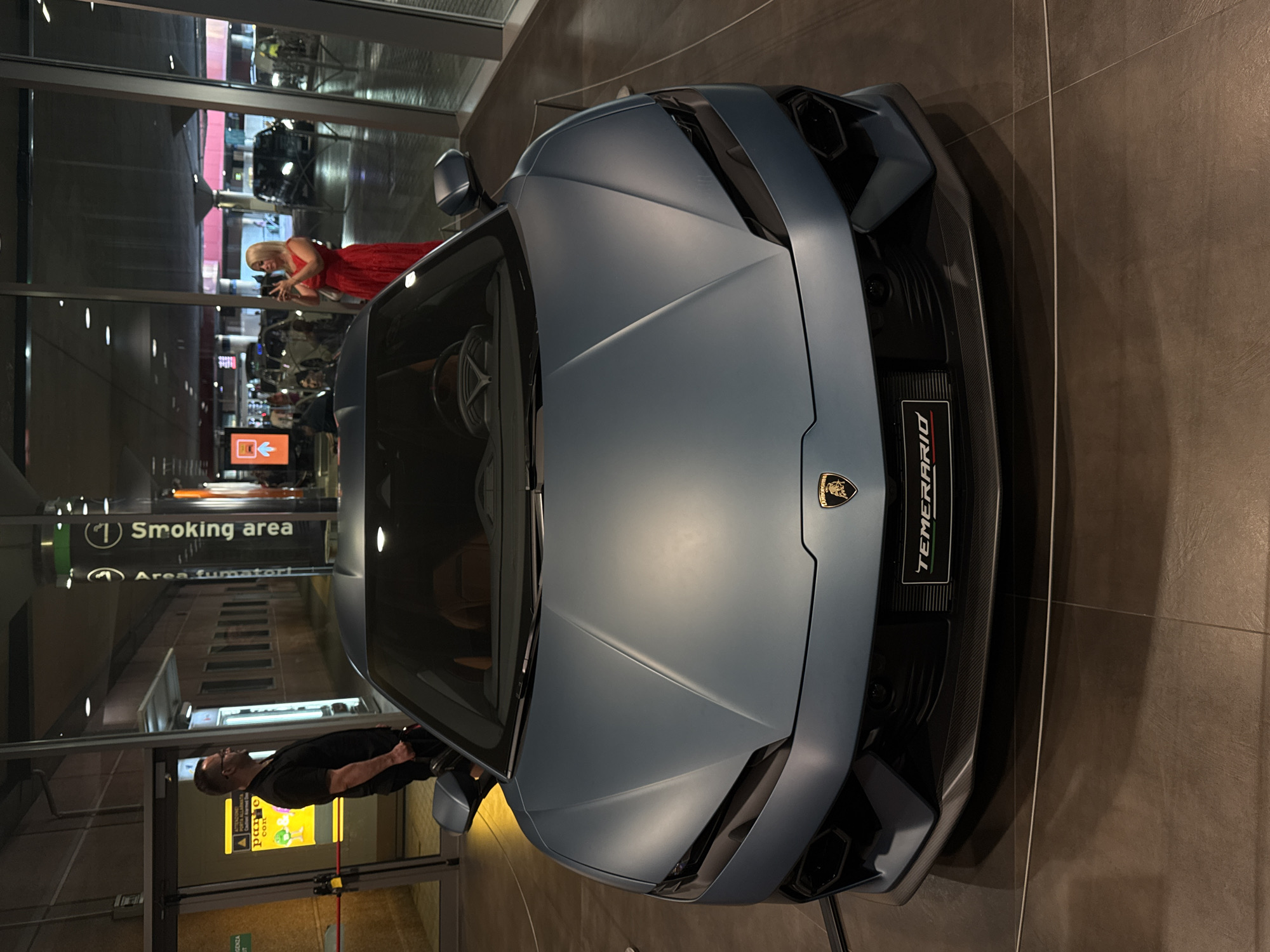 The image shows a matte blue Lamborghini Urus parked indoors. A sign below the car's front grille reads "TEMERARIO." In the background, through a glass window, a woman in a red dress is taking a photo with her phone. To the left, a man in a black shirt stands near a doorway, holding a black bag. The location appears to be a modern building with tiled floors and large glass windows. The scene is consistent with a showroom or an exhibition space within Bologna, Italy, given the car's marque and the typical modern architectural style found in such venues within the city.