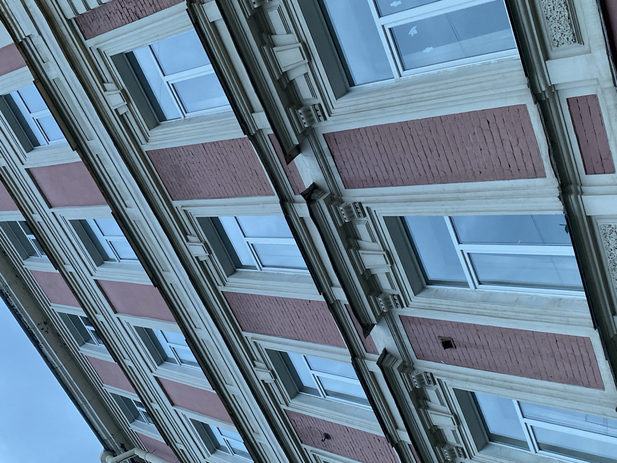 The image shows the exterior of a multi-story building, likely in Saint Petersburg, Russia, based on the architectural style. The building features a repetitive pattern of windows with light-colored frames and reddish-brown brickwork between them. The windows reflect the sky, suggesting daylight. The architecture includes decorative moldings and elements between each window section. The photo is taken from a low angle, looking upwards, creating a distorted perspective.