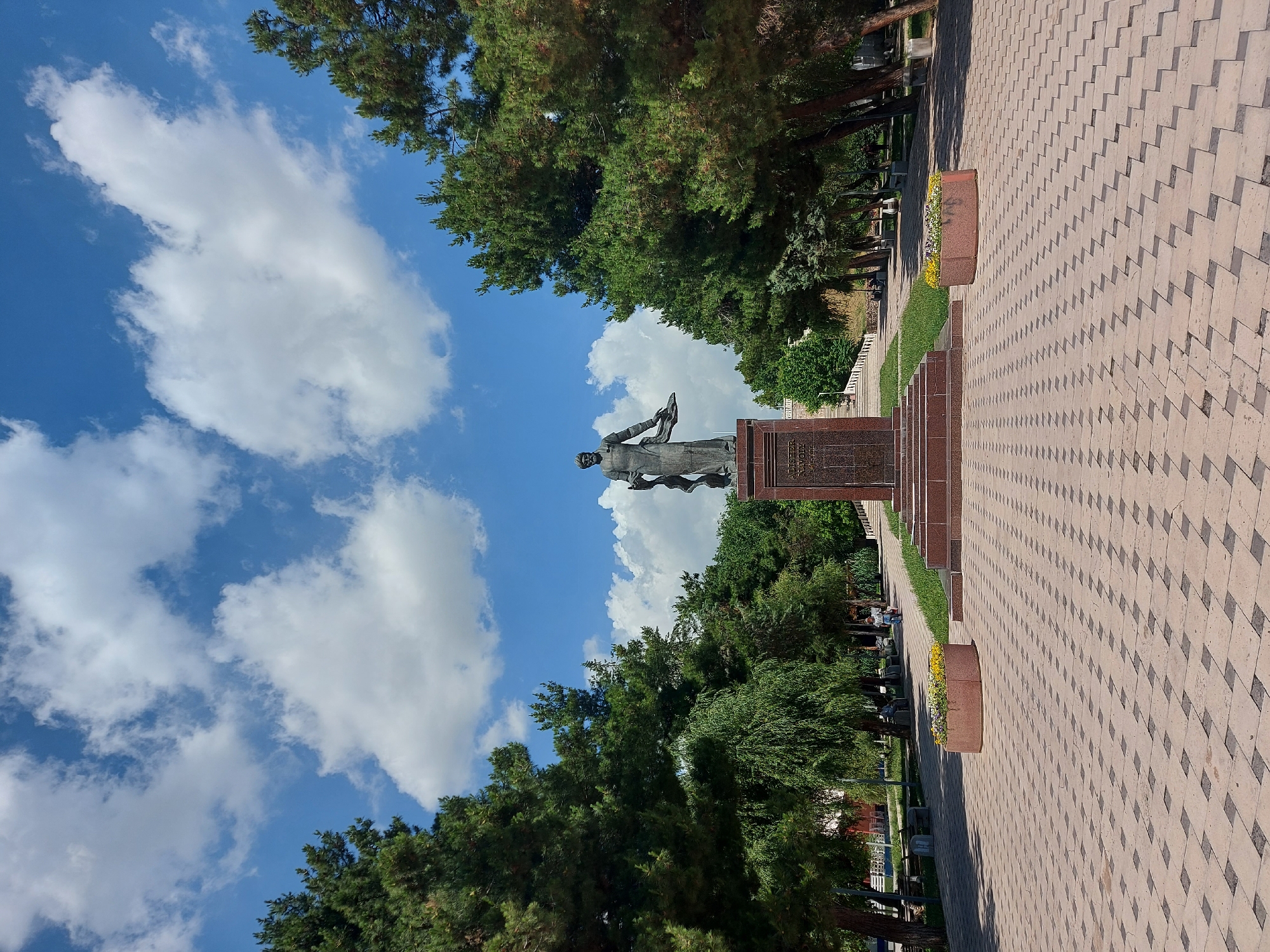 The image shows a statue in Tavaksay, Uzbekistan, with a sky of blue and puffy white clouds in the background. The statue is on a raised base of stone blocks. Trees surround the statue and the pathway. The pathway is made of light and dark rectangular pavers, and has flowerbeds along the sides.