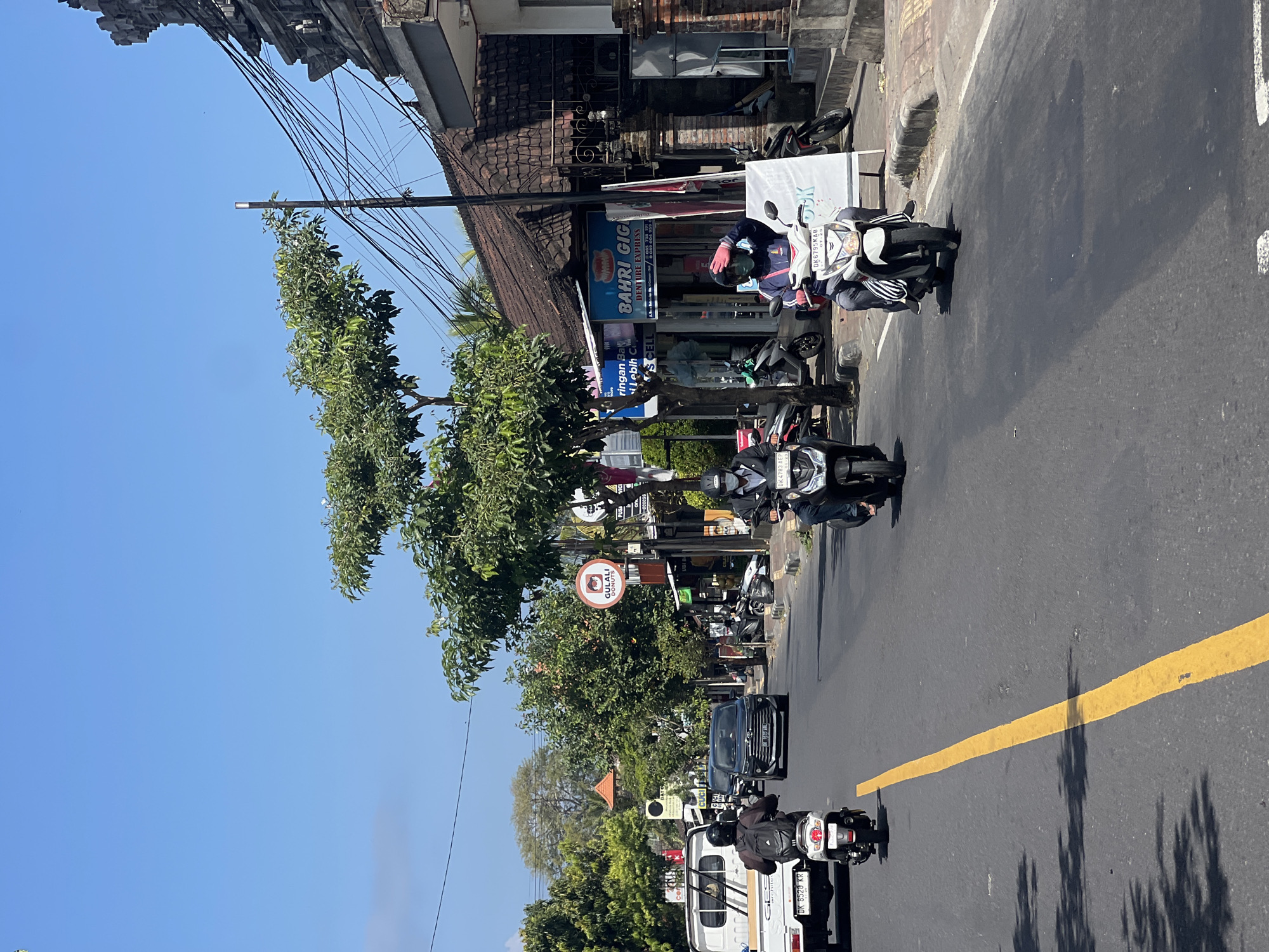 This image captures a street scene in Sukawati, Indonesia, on a clear, sunny day. The foreground features a paved road with a prominent yellow line dividing the lanes. Several individuals are riding motorcycles, with one prominently positioned in the center, wearing a blue jacket, mask, and pink gloves. Another motorcyclist is seen on the right, wearing a helmet and face mask, and a third is on the left, also wearing a helmet and backpack. Cars and other motorcycles are visible further down the road. The background reveals a row of shops and buildings with traditional tiled roofs, adorned with various signs, including one for "BAHRI GIGI DENTURE EXPRESS." Lush green trees and utility poles with a complex web of wires line the street, contributing to the tropical atmosphere. The sky is a bright, clear blue.