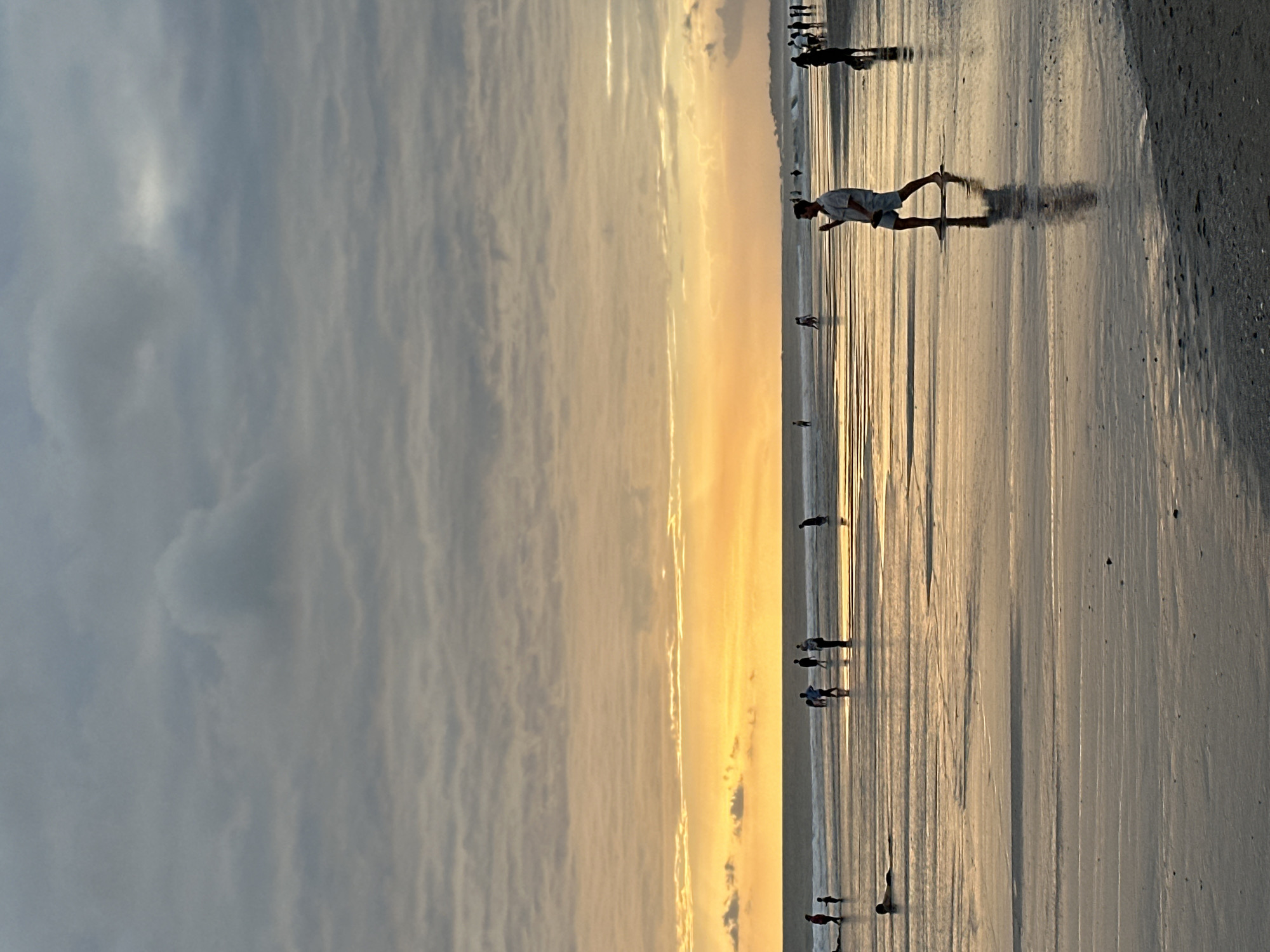 This media file captures a serene beach scene at sunset in North Kuta, Indonesia. The primary subjects are silhouettes of people walking along the wet sand near the shoreline. The image is oriented vertically, with the sky taking up the left half and the beach and ocean filling the right.
The sky is characterized by a gradient of colors, transitioning from a dusky grey with wispy clouds on the left to a warm, golden hue as it meets the horizon, indicating sunset. The low angle of the sun casts long shadows of the people onto the wet sand, creating a dramatic visual effect. The ocean is calm, with gentle waves lapping at the shore, reflecting the warm light of the sunset.
Several individuals and small groups are visible scattered across the beach. One person in the foreground is captured mid-stride, their shadow stretching out behind them. Others appear to be walking leisurely, enjoying the end of the day. The overall atmosphere is peaceful and tranquil, conveying a sense of calm and relaxation. The wet sand, the reflections, and the soft lighting all contribute to this mood. There is no visible text in the image.