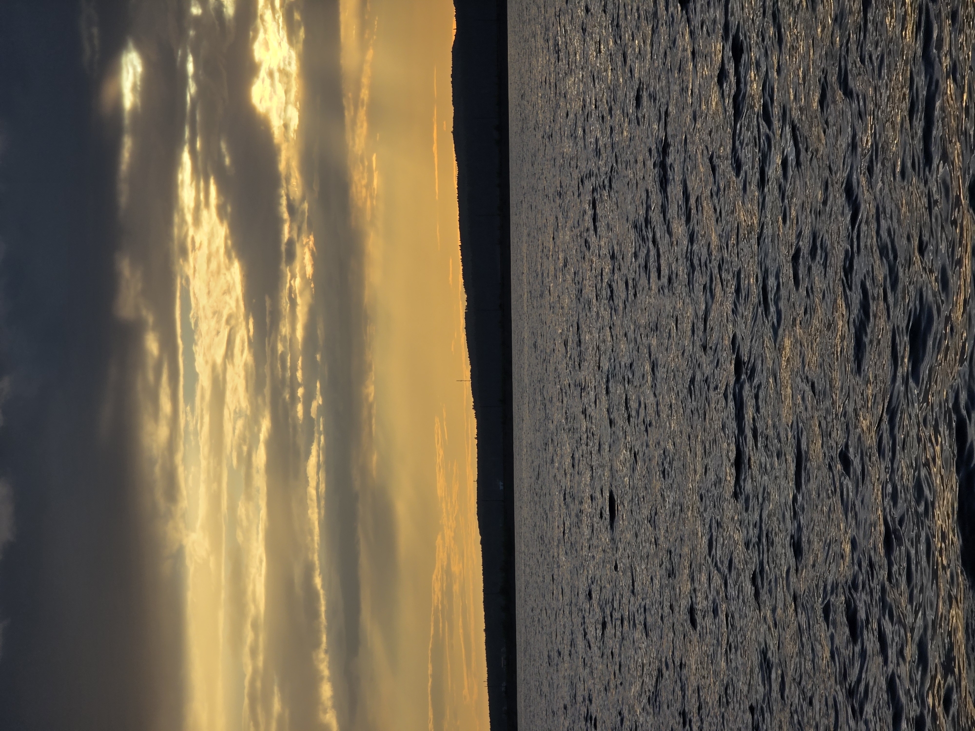 The image depicts a sunset over a body of water, likely the Kama River, in Chaikovsky, Russia. The sky is filled with dramatic clouds, ranging from dark gray to vibrant golden hues as the sun sets. Rays of light pierce through the clouds, illuminating the horizon. In the distance, a silhouetted treeline forms the edge of the land, with hints of distant structures visible. The foreground is dominated by the dark, rippling surface of the water, reflecting the colors of the sky. There are no people or specific objects of note in the scene. The overall atmosphere is serene and natural, characteristic of a sunset by a large river.