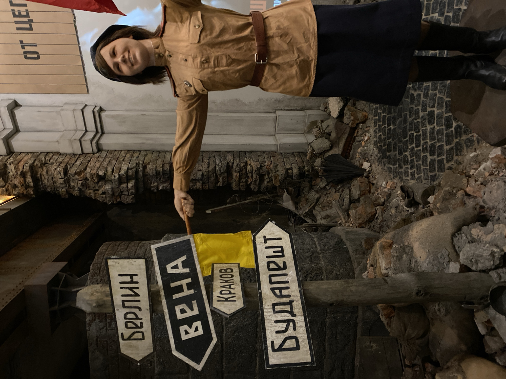 A museum exhibit, likely in Moscow, Russia, portrays a female reenactor dressed in a military uniform. She stands adjacent to directional signs pointing towards Berlin, Vienna, Krakow, and Budapest. The setting suggests a post-conflict scene amidst simulated rubble. The woman holds a pointer or stick. The overall impression is that of a historical or WWII-themed exhibit.