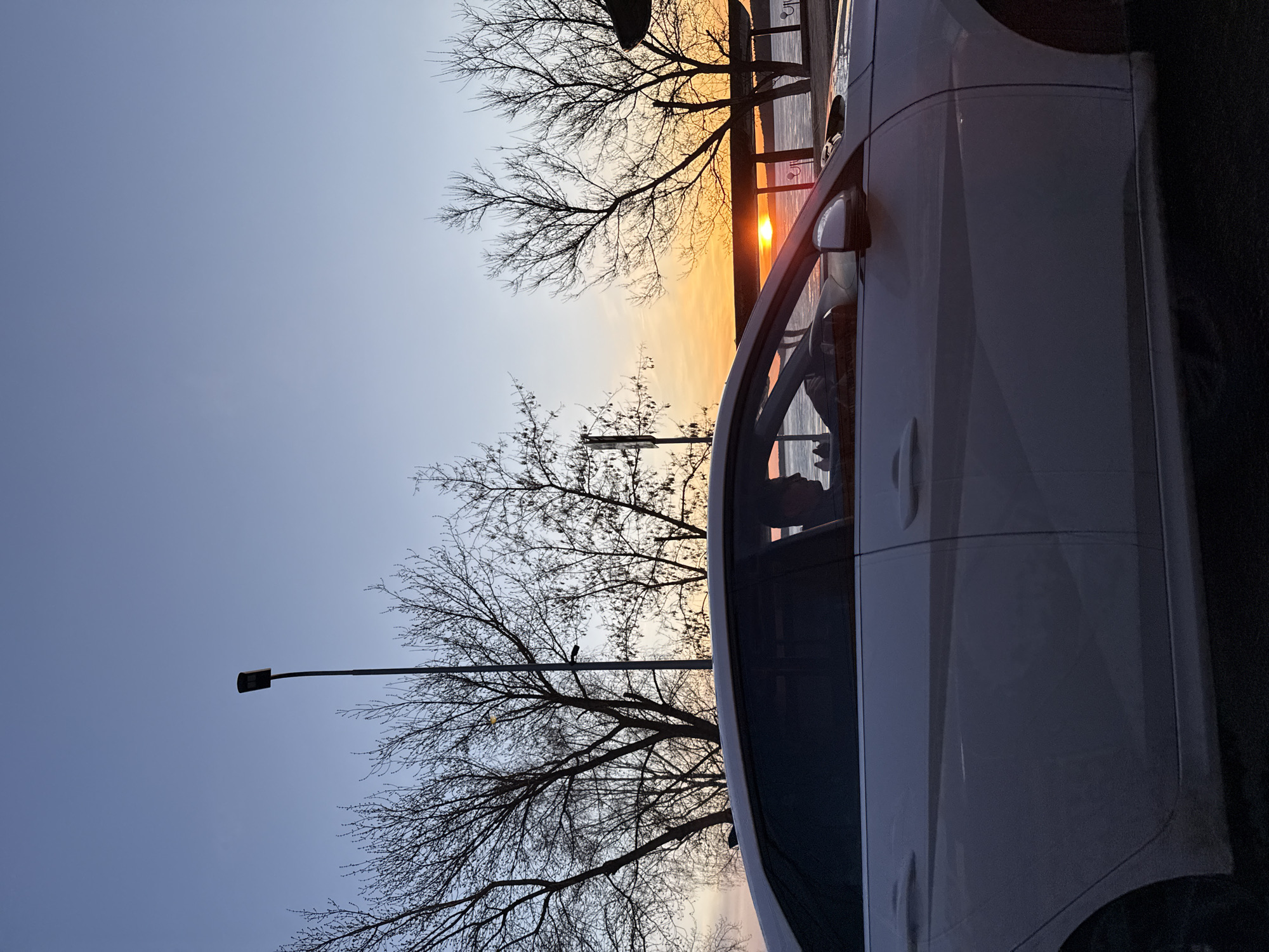 The image shows a partial view of a white car parked on a street in Votkinsk, Russia. The car is positioned on the right side of the frame. In the background, bare trees are silhouetted against a sky with a gradient of colors, suggesting either sunrise or sunset. A lamppost is visible on the left, near the trees. The sun is partially obscured by a building structure, with a bright glare indicating its presence. The environment suggests an urban or suburban setting during either early morning or late evening.