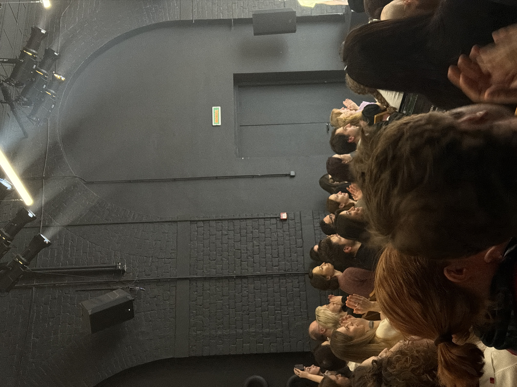 This high-angle image captures a crowd of people gathered in a dark, indoor performance venue, likely a concert hall or club, viewed from above looking down on their heads and shoulders. The attendees are facing forward, presumably towards a stage or focal point, and several individuals are seen clapping, indicating engagement with an event.
The venue features dark grey or black walls, with sections of smooth finish and other areas showcasing exposed brickwork, suggesting an industrial or renovated aesthetic. On the left side of the image, multiple stage lights or spotlights are mounted on a structure, casting bright beams. Two rectangular speakers are visible, mounted on the walls. An arched architectural element is also discernible on the left.
Visible text includes a green sign on a smooth wall section that reads "ВЫХОД" (VYKHOD), which is Russian for "EXIT." Another partial text, "GHT SKY," is faintly visible near the lighting equipment. A small, indistinct red sign is also present on a brick wall section. The overall lighting is dim, characteristic of a performance space, with highlights provided by the stage equipment. The scene depicts an active event with an appreciative audience in Moscow, Russia.