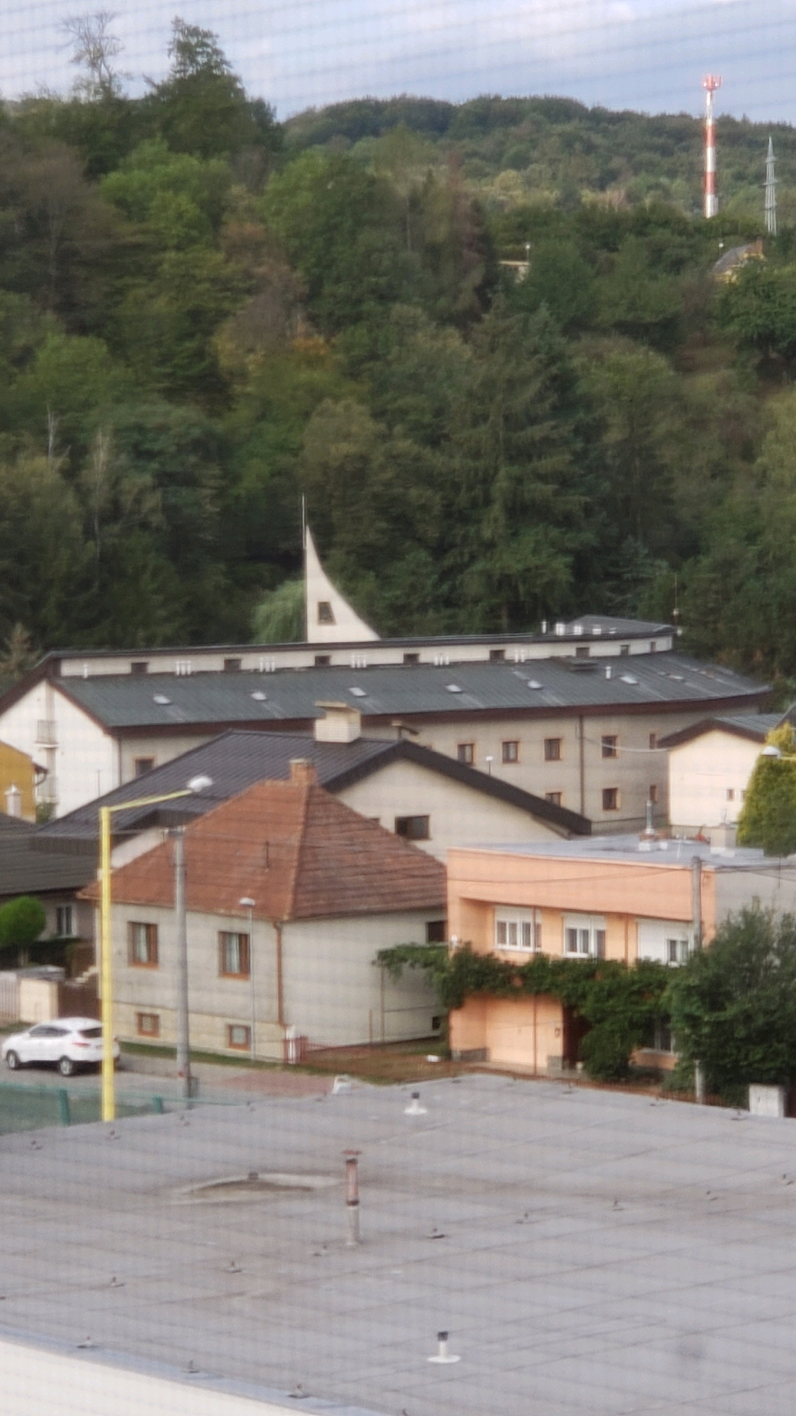 The image shows an exterior view of buildings in Prešov, Slovakia, with a wooded hillside in the background. In the foreground, a flat, grey rooftop with several ventilation pipes and anchor points is visible. Behind this rooftop, there are several buildings of varying heights and architectural styles.
To the left, a building with a light-colored facade and a brown tiled roof is prominent. It has several windows and a chimney. A bright yellow pole, possibly a street light, stands in front of this building. A white car is parked on a paved area to the left of this building.
Adjacent to the brown-tiled roof building, a larger, modern building with a dark grey, angled roof and multiple skylights dominates the mid-ground. This building has a light-colored facade with numerous windows and appears to be a multi-story structure.
Further to the right, a lighter orange-colored building with white-framed windows is visible, partly obscured by greenery.
In the background, the wooded hillside rises, covered in dense green trees. A tall, slender white and red striped tower, likely a communication tower, is visible on the crest of the hill, along with a more distant, darker tower structure. A distinctive triangular architectural feature, possibly part of a church or other public building, can be seen emerging from behind the modern grey-roofed building. The sky above is overcast.