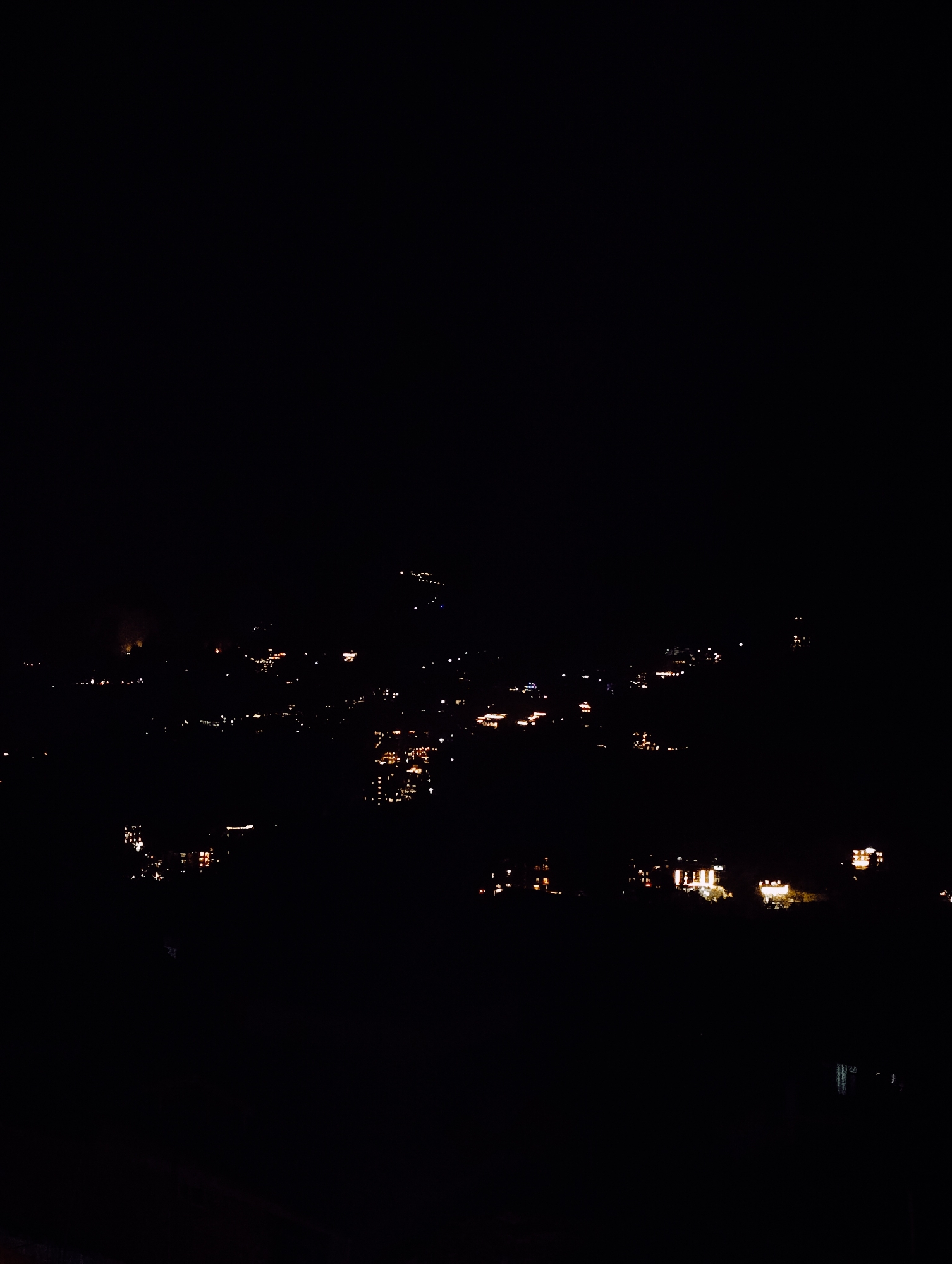 The image depicts a nighttime aerial view of a cityscape, likely Vashisht, India, characterized by the glow of scattered lights against the dark expanse. The lights suggest the presence of buildings and possibly streets, illuminating portions of the otherwise obscured landscape. The scene is dominated by darkness, with the artificial lights serving as the primary visual elements, creating a sense of depth and scale.