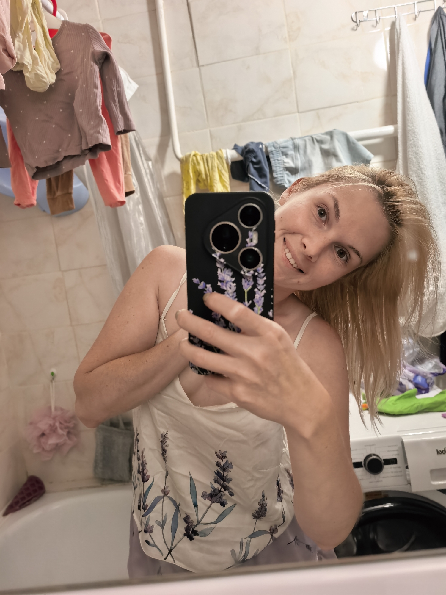 The image is a selfie taken in a bathroom, likely in Sterlitamak, Russia, as indicated by the user prompt. A blonde woman is looking into a mirror, holding a smartphone. She is wearing a white camisole with a floral pattern. In the background, clothes are hanging on a rack, a washing machine is visible, and a bathtub is present. The bathroom appears to be functional and lived-in.