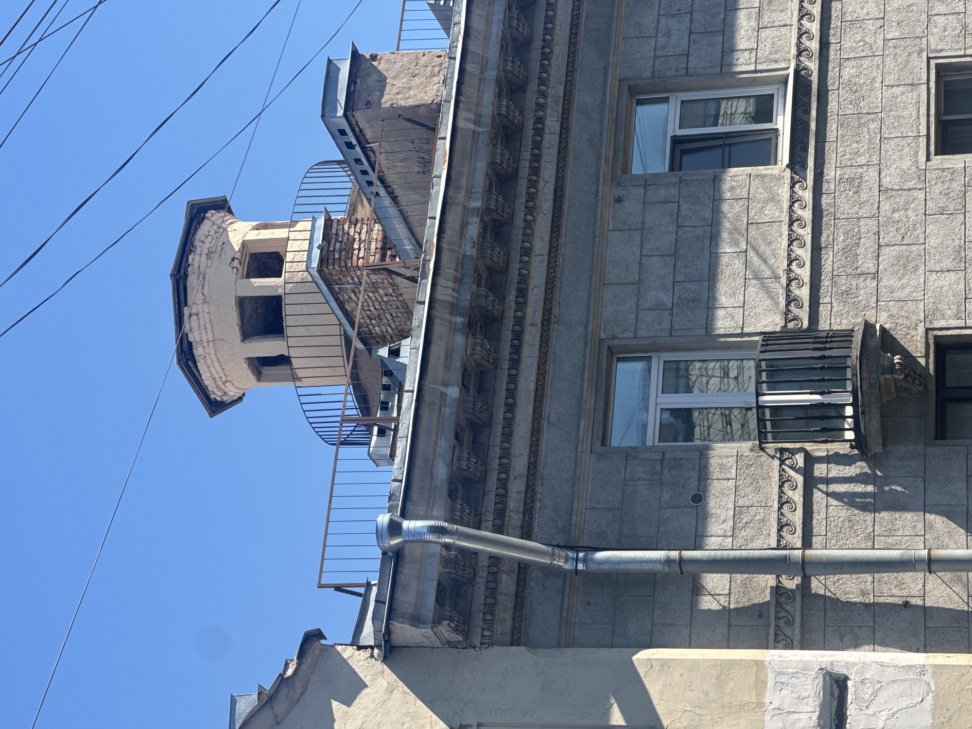 This is a vertical, eye-level shot of the exterior of a building on a clear, sunny day. The building appears to be old and possibly in disrepair, with exposed brick and crumbling plaster visible in some areas. Electrical wires crisscross the bright blue sky above.
The main subject is a section of the building's facade, constructed of large, square, grey stones. There are at least two windows visible, both with white frames and clear glass. One window has a decorative metal grate in front of it. A metallic downspout runs horizontally across the building, attached by bands. Above this, a section of the building features ornate architectural detailing, including decorative moldings and what appear to be carved floral motifs.
To the left of the main facade, a more deteriorated section of the building rises. It has a rounded, turret-like structure with openings that reveal a brick interior. A metal fire escape or exterior staircase with a curved railing is attached to this section and extends downwards.
The sunlight casts strong shadows on the building, suggesting it is mid-day. The overall impression is one of an urban environment, possibly in an older part of a city, with a mix of architectural styles and signs of aging. No people are visible, and no text is discernable in the image.
