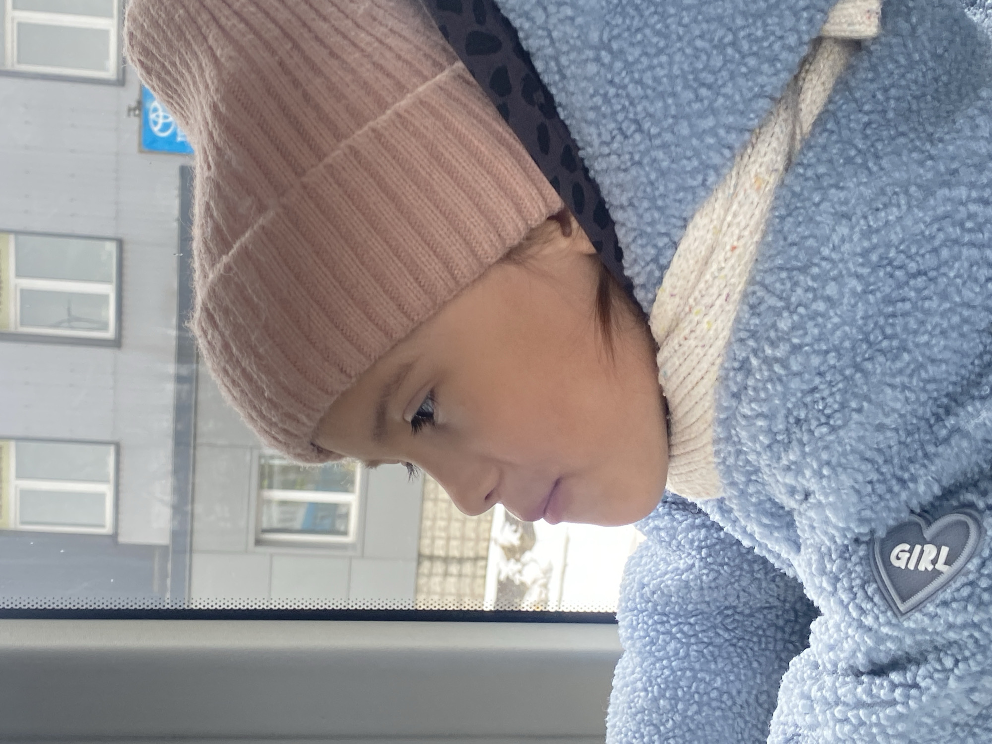A young girl in a pink beanie and a blue fleece jacket is looking downward, potentially out of a window. The backdrop shows a multi-story building, and a sign indicates a location in Chaikovsky, Russia.
