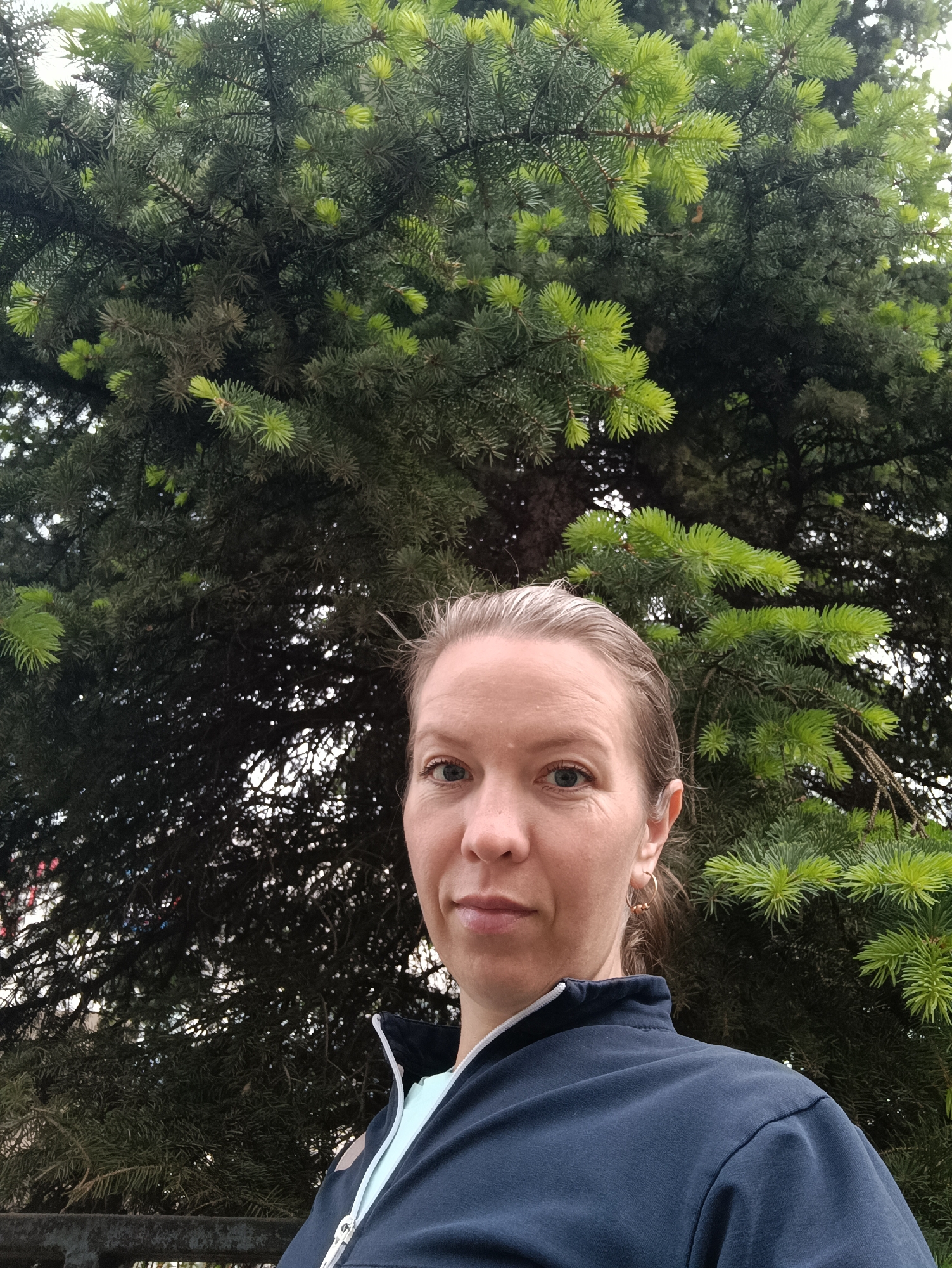 A woman is standing outdoors in front of a large evergreen tree. The tree has dense, green needles and some lighter green new growth at the tips of its branches. The woman is fair-skinned with blue eyes, and her hair is pulled back. She is wearing a dark blue jacket with a white zipper and a light blue shirt underneath. She is wearing gold hoop earrings. The photo appears to be taken during the daytime with natural lighting. Based on the context provided and assuming that evergreen trees of this type are common in the city of Chaikovsky, Russia, it is plausible that the photo was taken in a park or natural area within that city.