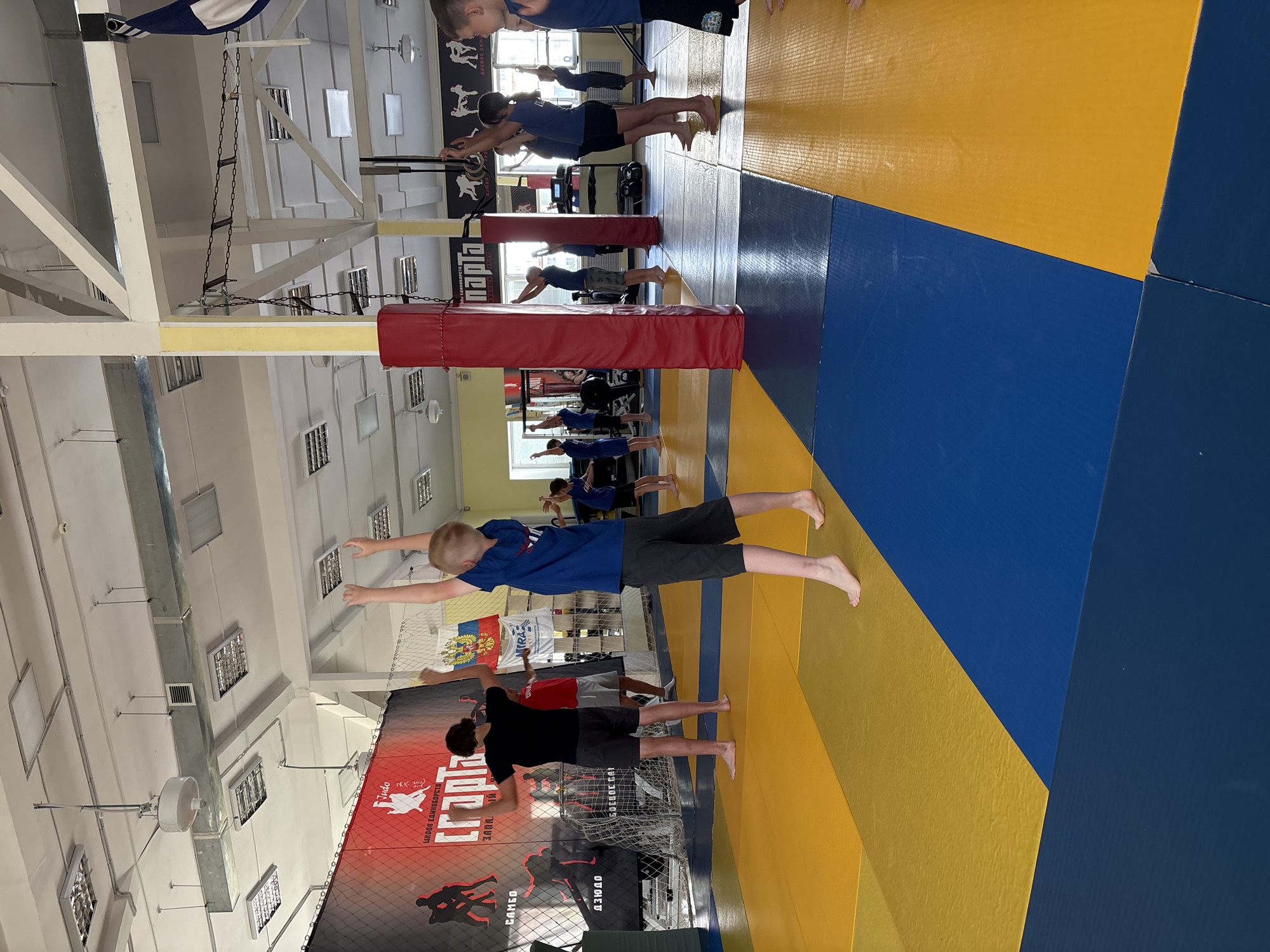 The image shows a group of children and adolescents engaged in a physical activity in a gym. The gym has a brightly colored mat on the floor, with alternating sections of yellow and blue. There are various pieces of sports equipment visible, including a climbing rope and some punching bags in the background. The walls are adorned with banners and logos, some of which appear to be related to martial arts or sports clubs. The ceiling is industrial, with exposed pipes and lighting fixtures. The overall impression is of a sports training facility. The scene is consistent with the Judo hall of the Chaikovsky Sports and Recreation Center "Neftekhimik" located in Chaikovsky, Russia.