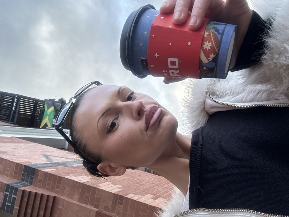 A woman in a black top and white furry jacket is holding a coffee cup. The cup is blue and red with a star design. Sunglasses are perched on her head. The background reveals a brick building with a Jamaican flag. The sky is overcast. The photo is taken from a low angle, looking up at the woman.
