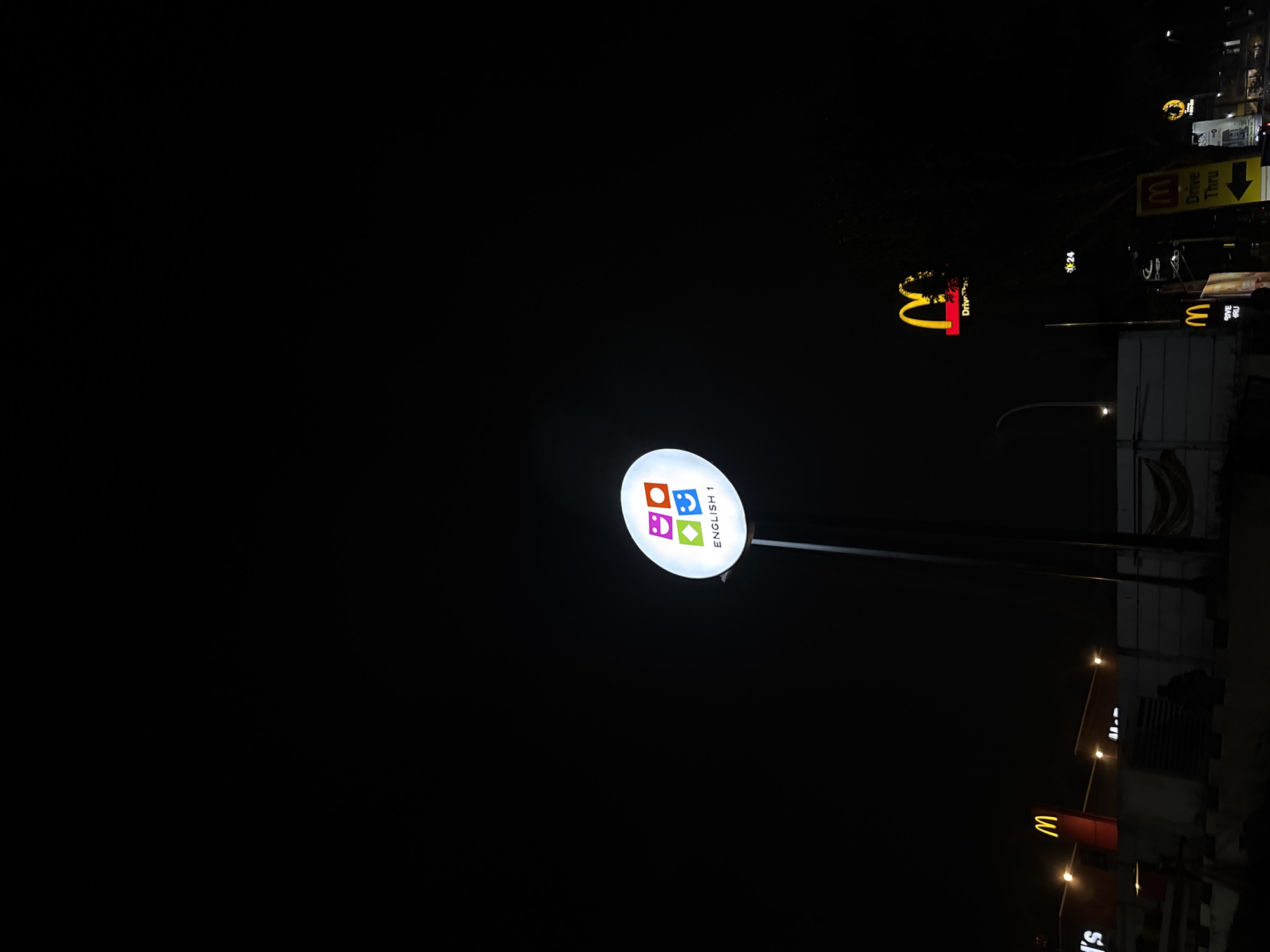 The image captures a nighttime scene in a commercial area of South Tangerang, Indonesia, illuminated by artificial light from business signs against a dark sky. No people are visible.
In the mid-ground, a prominent, circular, white illuminated sign stands on a pole. This sign features a logo composed of four colorful square icons—orange, red, blue, and green—each containing a white symbol resembling a simple face or emoji. Below these icons, the text "ENGLISH 1" is clearly displayed in black.
In the background, multiple illuminated signs for McDonald's are visible, primarily to the right. One large McDonald's sign features the iconic yellow "M" arch logo, with the text "Drive Thru" visible beneath it. Another smaller McDonald's sign, also displaying the "M" logo, is seen further back, with the number "24" indicating 24-hour service. Other faint lights and indistinct building structures are discernible in the distance, suggesting a typical urban commercial setting after dark. The clear night sky is dark, providing a stark contrast to the brightly lit signs.