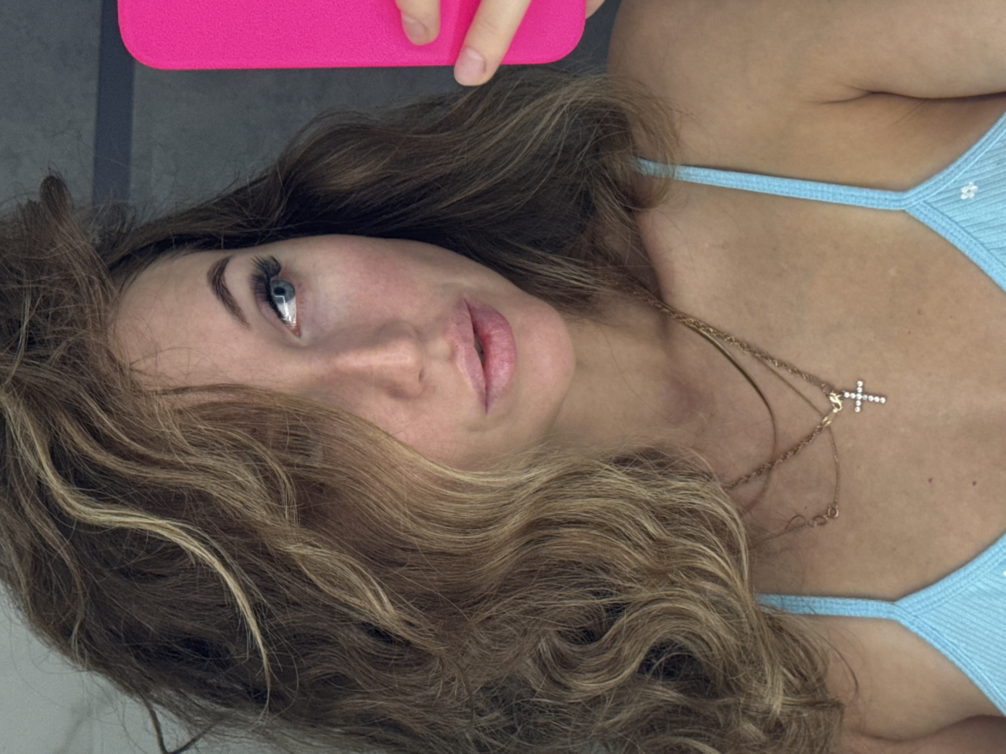 A rotated close-up selfie captures a young woman from an overhead perspective, appearing to lie down. She holds a bright pink smartphone, partially visible in the upper left, with her fingers grasping its edge. Her long, wavy, light brown hair with blonde highlights frames her face, fanning out around her head.
Her blue left eye is prominent, featuring long, dark eyelashes and a well-defined eyebrow. Her lips are slightly parted, showing a soft pink lip color, and her expression is neutral to gently pensive. She wears a light blue, ribbed top or bikini top with delicate straps and small white floral patterns. A gold-colored layered necklace adorns her neck, featuring a sparkling cross pendant and another smaller pendant.
The background is a plain, dark grey surface, indicative of an indoor setting. The image's orientation suggests she is lying on her side or the phone was held rotated during capture. No visible text is present. This scene is set in Chaikovsky, Russia.