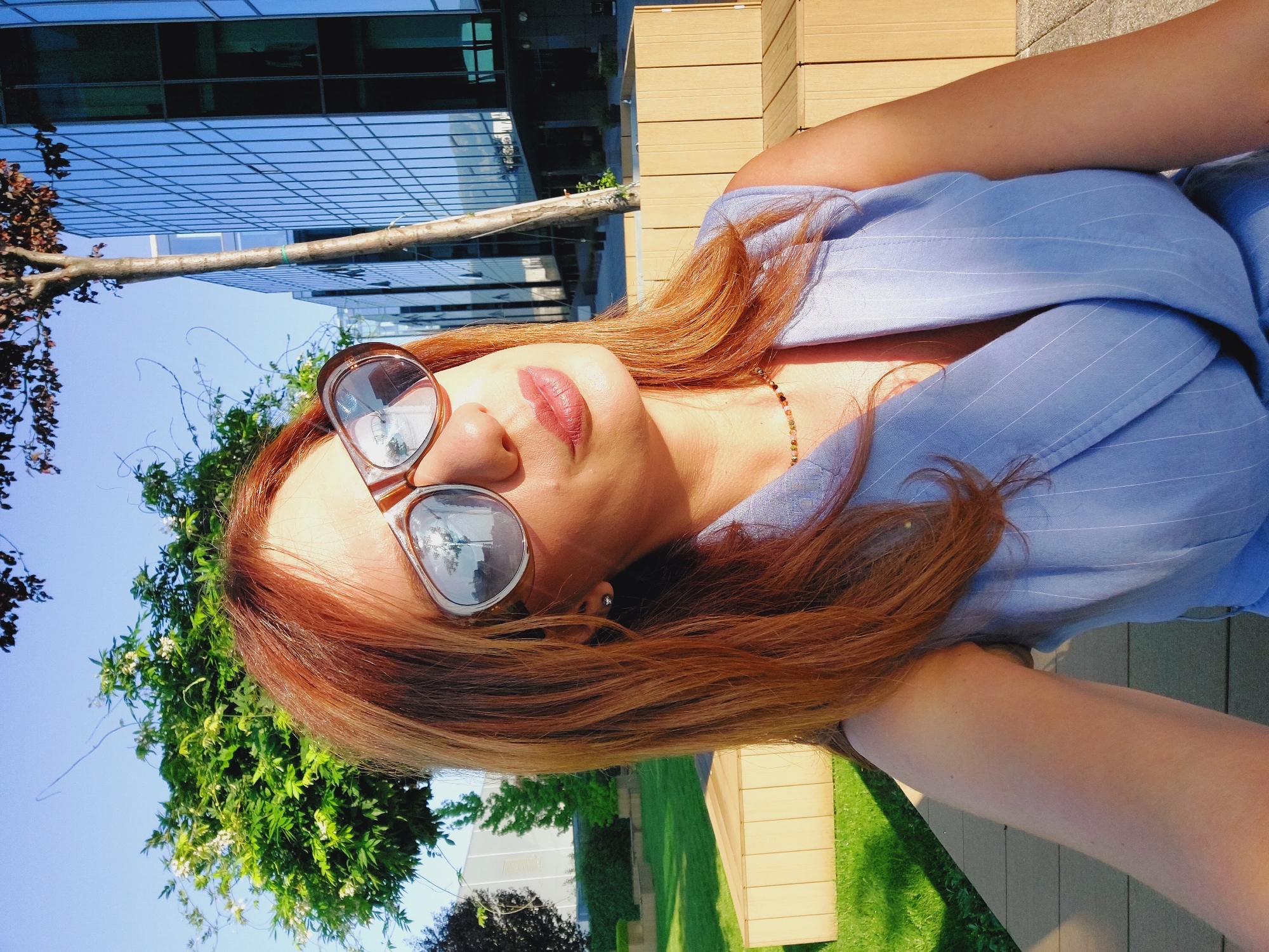 A woman with auburn hair and sunglasses is posing for a selfie outdoors, with a modern office building and greenery in the background. The building features a facade of reflective glass, and a tree is planted in front of it. The woman is wearing a light blue pinstriped sleeveless top and a beaded necklace. She is seated on a wooden bench in a landscaped area with a green lawn. The scene suggests a sunny day in an urban environment, possibly a business district. The location is not specified as Sofia, Bulgaria.