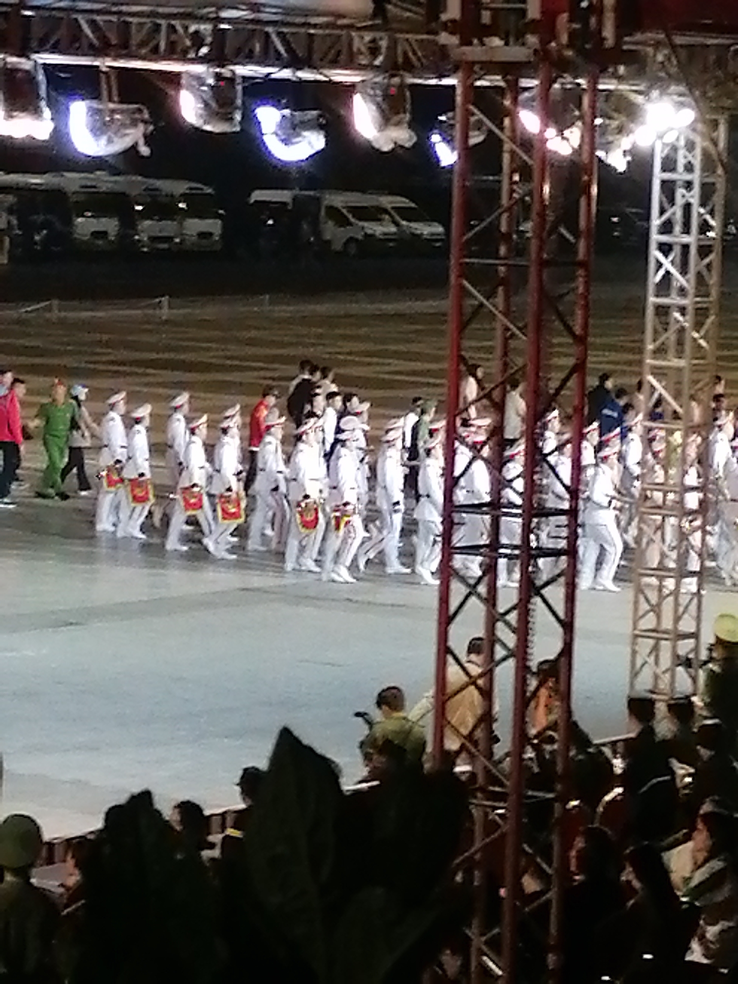 The image depicts an outdoor event at night, likely a parade or a ceremony, set against a backdrop of bright stage lights and scaffolding. In the foreground, a crowd of spectators is visible, silhouetted against the illuminated performance area. The main subjects are a group of people in white uniforms with red sashes, marching in formation. They are wearing white hats and appear to be carrying drums, suggesting a marching band or ceremonial procession.
Behind the performers, several white vans or buses are parked in rows, partially illuminated by the stage lights. The ground appears to be a light-colored, smooth surface, possibly a stage or a specially prepared area for the event. The overall atmosphere is festive and organized, with the lighting creating a dramatic effect. The presence of stage lights and scaffolding indicates a prepared venue for a public performance. The time of day is clearly night due to the artificial lighting and the dark surroundings. While the specific location is Da Lat, Vietnam, there are no overt visual cues to specifically identify this. The emotions of the spectators are not discernible due to the silhouettes, but the organized nature of the event suggests a formal or celebratory occasion. No visible text is present in the image.