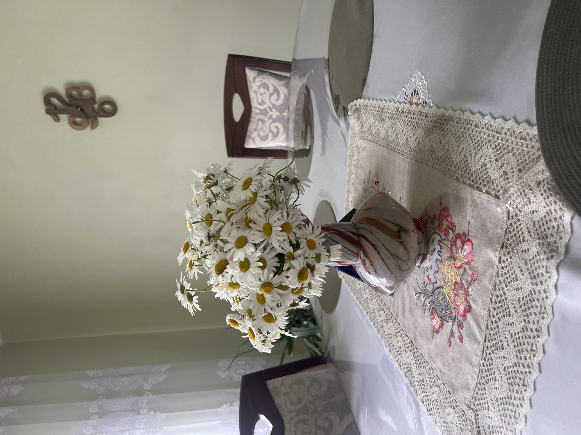 A bouquet of white daisies in a multi-colored vase sits on a table covered with a clear plastic tablecloth and a crocheted doily with floral embroidery. Two chairs with upholstered seats are visible on either side of the table. A wooden clock is mounted on a pale green wall in the background. The scene appears to be an interior dining area in a home or a small establishment. No people are present. The lighting suggests it is daytime, possibly with natural light coming from a window off-camera to the left.