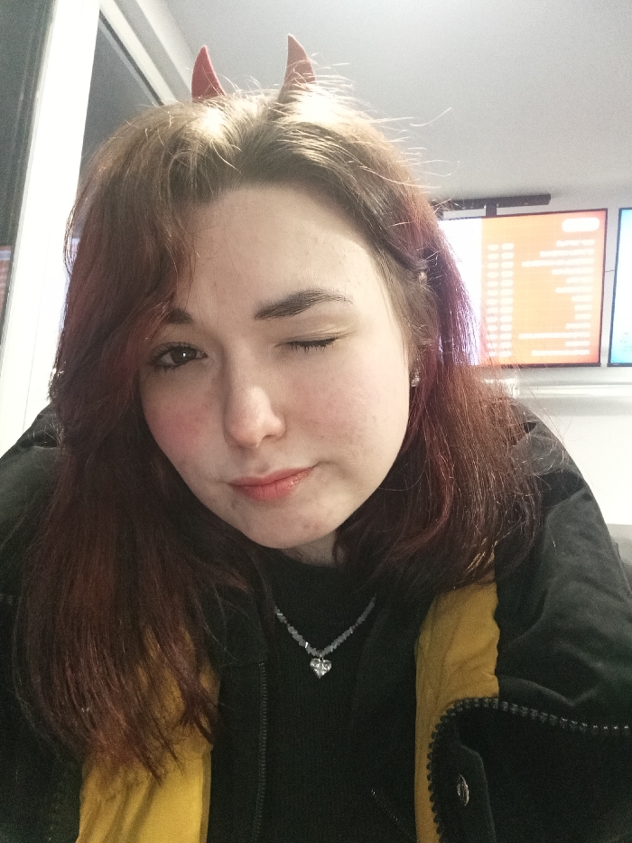 A fair-skinned woman with auburn hair winks at the camera. Small, red devil horns adorn her head. She wears a black sweater, partially covered by a black and yellow jacket. A delicate necklace with a heart pendant is visible. In the background, a brightly lit room contains a window and a double screen displaying text and graphics. The overall impression is casual and playful.