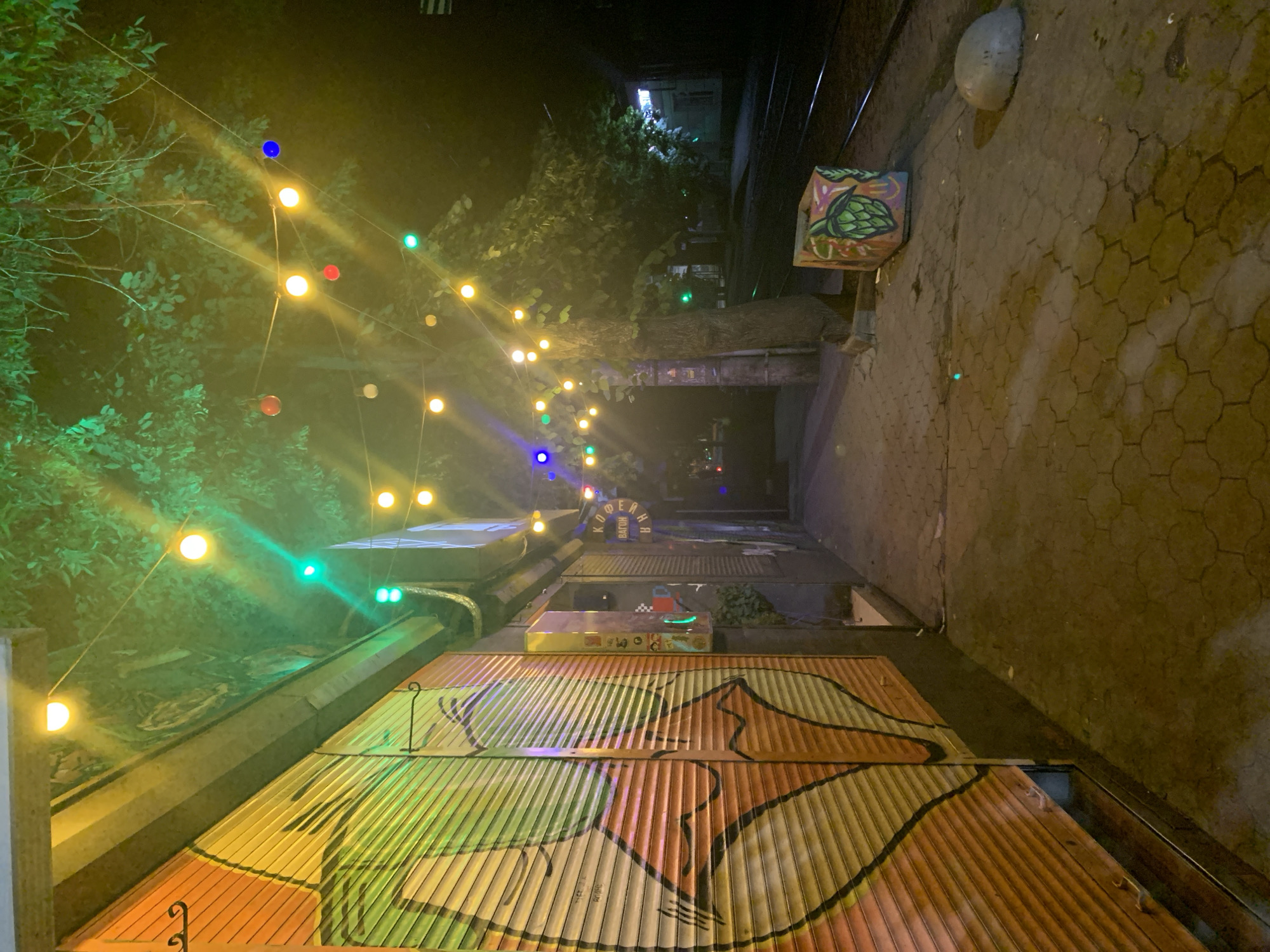 An eye-level, medium shot captures a dimly lit urban alleyway at night, illuminated by strings of colorful lights strung between trees and buildings. The left side of the frame is dominated by a large, rolling metal security gate covered in vibrant, graffiti-style artwork featuring abstract shapes in orange, yellow, and green. To the right of the gate, the alley opens up, revealing a paved walkway with an uneven, textured surface. A tree with lush green foliage is visible on the right side of the alley, its branches draped with more colorful lights in red, blue, yellow, and green. Further down the alley, what appears to be a building facade with more graffiti and signage is visible. In the foreground, to the right, a concrete planter box decorated with a colorful image of hops sits on the pavement. Beyond that, railway tracks run parallel to the alley. A rounded concrete bollard is positioned on the walkway in the lower right corner. The overall atmosphere is vibrant and slightly mysterious, with the interplay of darkness and colorful illumination.