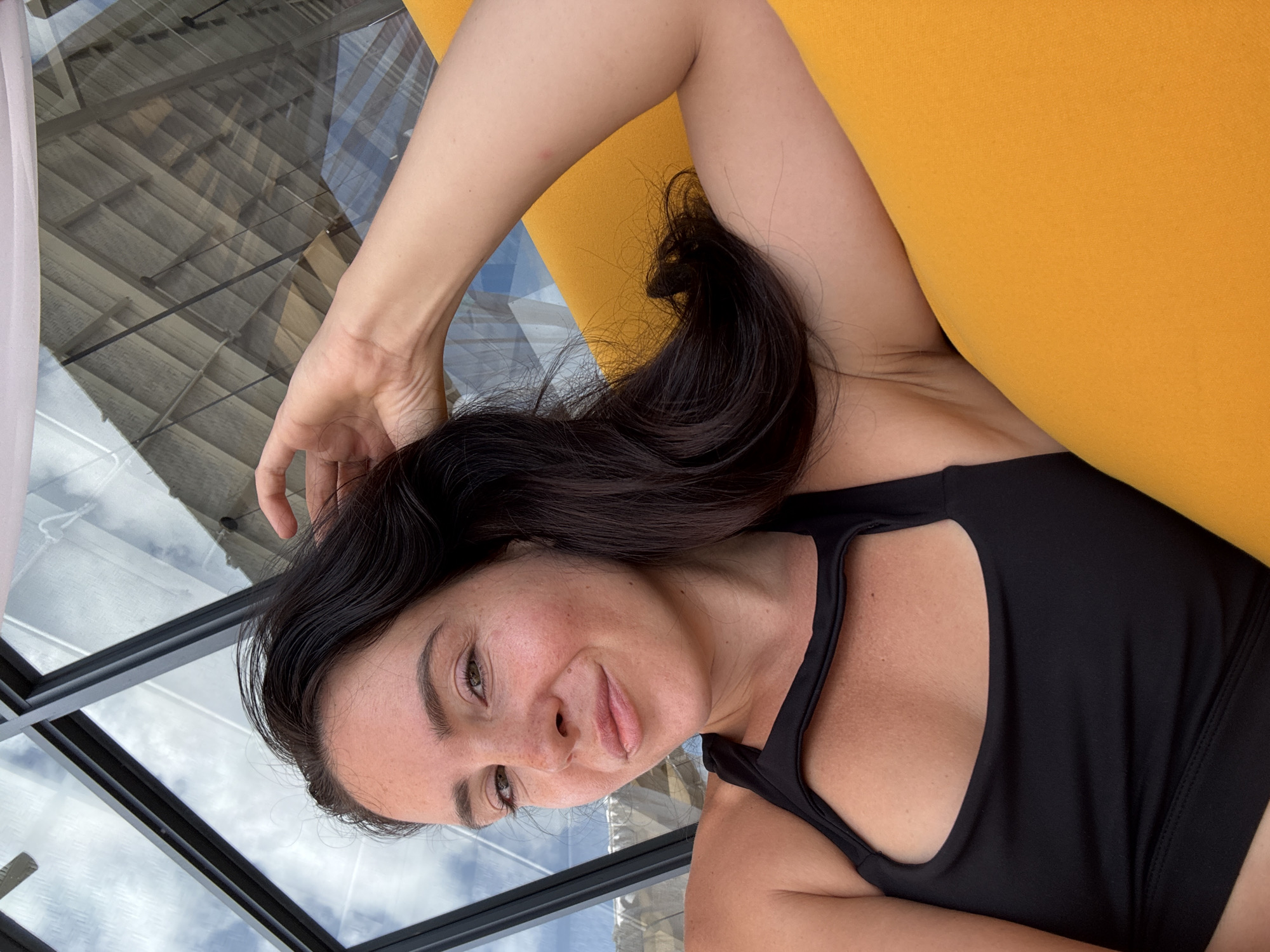 A dark-haired woman with freckles smiles at the camera, her right hand resting on her head. She wears a black sports bra with a cut-out detail and is reclined on an orange surface, possibly a lounge chair. The background features a glass ceiling with a view of the sky and a patterned ceiling structure. The overall setting suggests an outdoor or semi-outdoor location with natural light.