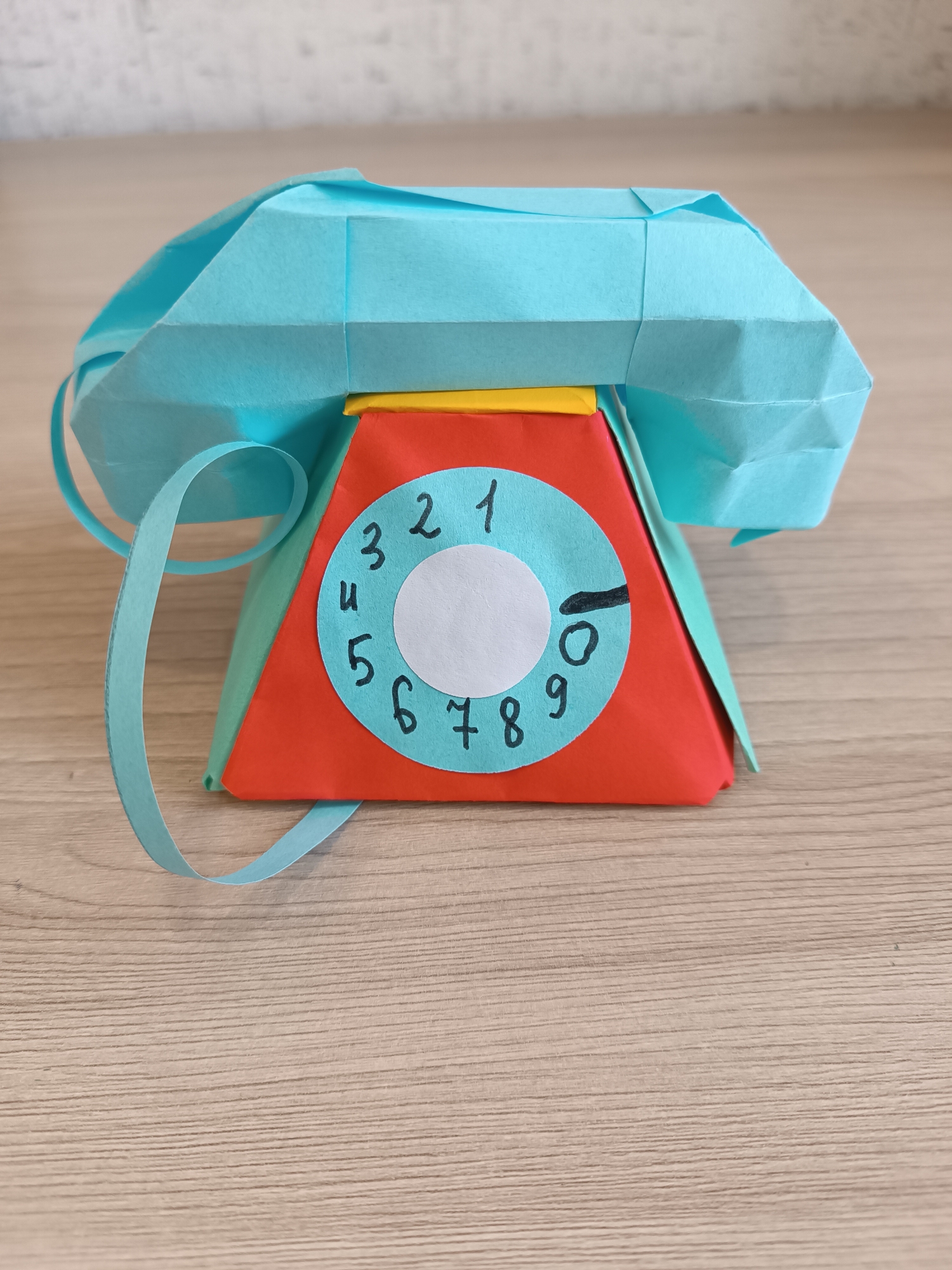 The scene depicts a handmade origami model of a vintage rotary telephone, situated on a light brown wooden surface with horizontal grain, in what appears to be an indoor setting in Chaikovsky, Russia. The telephone model features a light blue folded paper receiver resting atop a base that is predominantly red on the front, with light green sides and a thin yellow strip across the top edge. A light blue circular paper dial is affixed to the front, displaying handwritten black numbers 1 through 0 arranged clockwise around a central white circle. A black marker is positioned at the '0' on the dial. A thin strip of light blue paper, fashioned to resemble a coiled cord, extends from the left side of the receiver, curving onto the wooden surface. The background consists of a light-colored, subtly textured wall. No individuals are present, and no actions or interactions are depicted.