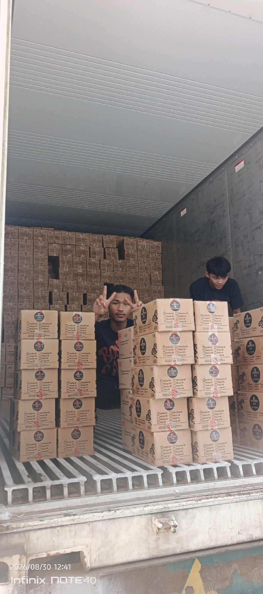 Two young men are inside the back of a truck filled with cardboard boxes. The boxes are stacked high and appear to be containing food items, possibly ice cream, as indicated by some of the labels on the boxes which include flavor names and quantities. The man in the foreground is making a peace sign with his fingers. The truck is parked on a surface that appears to be asphalt or concrete. The environment suggests a loading dock or a similar industrial area. The image has a timestamp in the bottom left corner indicating August 30, 2026, at 12:41 PM, and the device "Infinix NOTE 40". The overall scene is that of a logistics operation, likely in the city of Patampanua, Indonesia, given the timestamp and context.