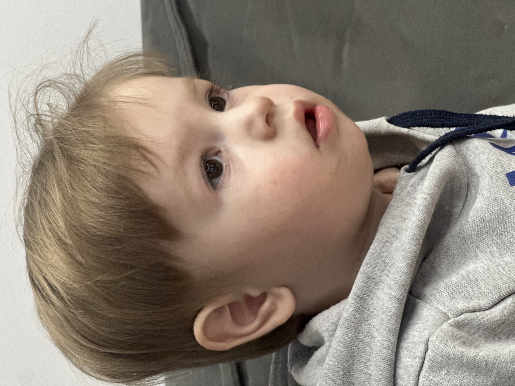 A close-up shot depicts a young child with fair skin and light brown hair. The child is lying down, wearing a gray hooded sweatshirt with a dark blue drawstring. The child's face is turned upwards and to the side, revealing a curious expression. The background is blurry, but a gray upholstered surface can be seen. The location is unknown; the information about Эстачи, Russia, is irrelevant.