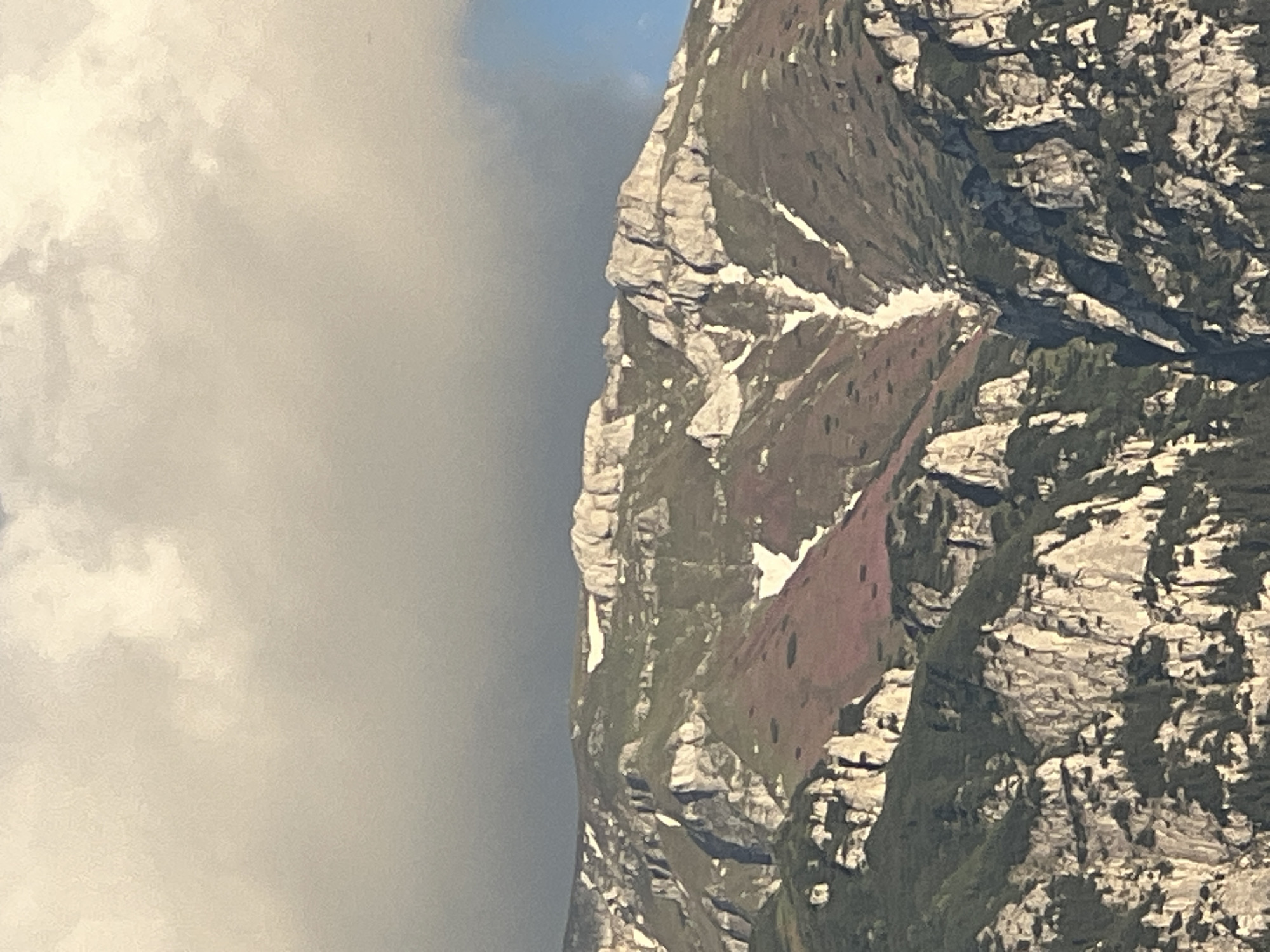 The image depicts a steep, rocky mountainside with patches of snow and vegetation. The mountain is textured with layers of rock and appears to be a rugged, alpine environment. On the left side of the image, a large expanse of cloudy sky is visible, with some brighter areas suggesting sunlight filtering through. The clouds are dense and diffuse, creating a soft, diffused light on the left portion of the frame. The mountain itself is in sharp focus, highlighting the granular details of the rock formations and the sparse green and reddish-brown plant life clinging to its slopes. Streaks of white snow are visible in crevices and on ledges, indicating a cool climate or high altitude. The lighting suggests it may be daytime, possibly with the sun partially obscured by clouds, as there are distinct shadows on the mountain. There is no visible text in the image. The scene evokes a sense of natural grandeur and the wildness of a mountainous landscape.
