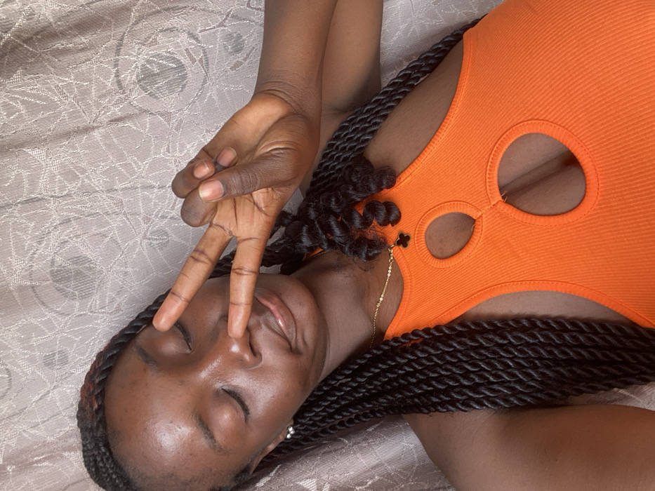 A dark-skinned woman lies on a patterned surface, likely a bed. She wears an orange top with cutouts. Her eyes are closed, and her hand is raised in a peace sign near her nose. She has long, dark braids framing her face. A thin gold necklace with a four-leaf clover charm is visible. The composition is an overhead shot.
