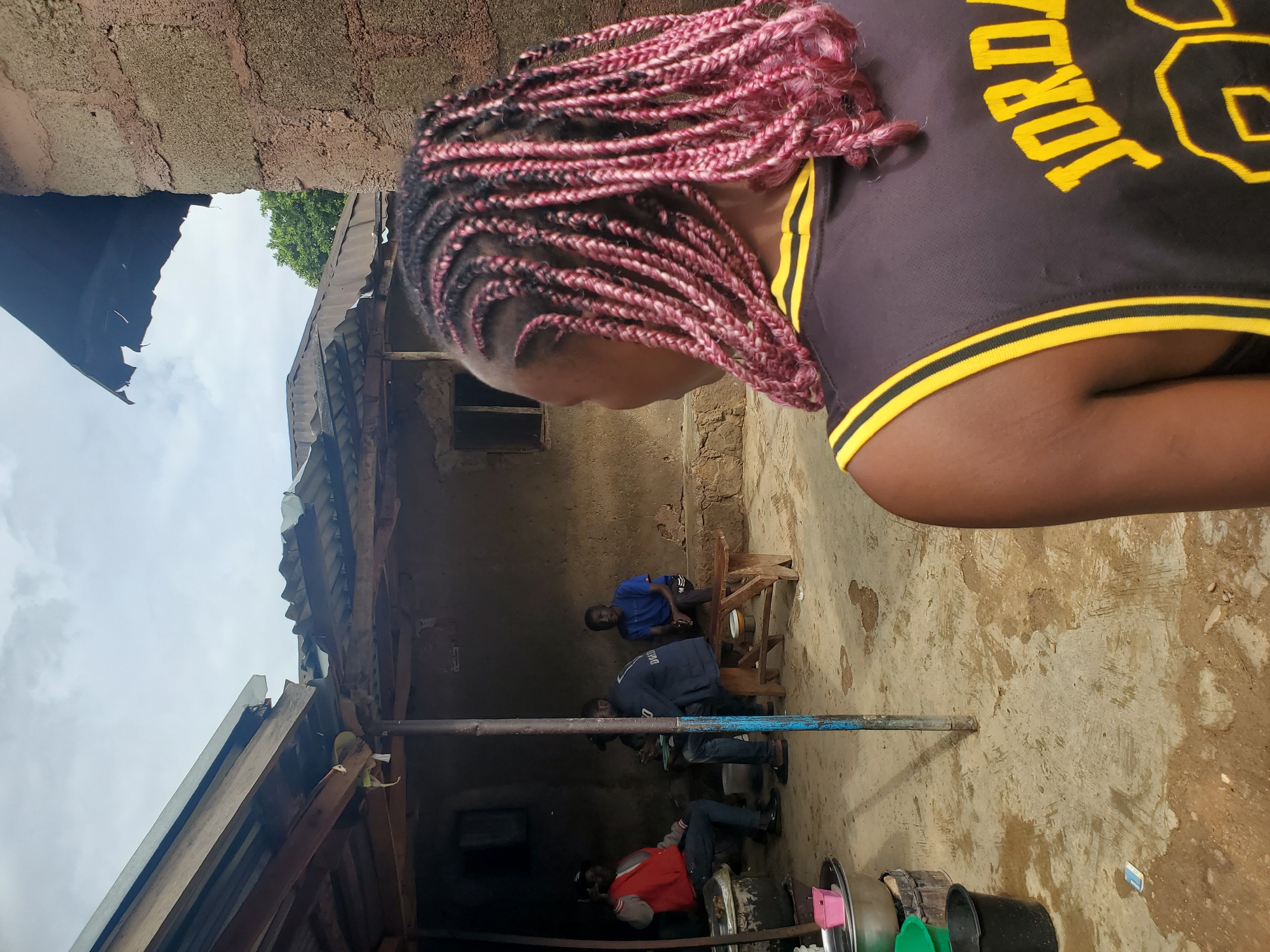 The media file depicts an outdoor scene in Jos, Nigeria, likely in a residential or communal area. The primary subject is a person from the shoulders up, facing downwards and to the left. They have long, braided hair with pink highlights. They are wearing a black t-shirt with yellow trim and yellow lettering that appears to spell out "JORDAN."
In the background, several other individuals are visible, seated and standing. One man in a blue shirt is on a wooden stool. Another man in a dark jacket is sitting on a similar stool. Further back, a person in a red jacket is also seated. The setting appears to be a courtyard or open space with a dirt or concrete floor. There are rudimentary structures around, including one with a corrugated metal roof, possibly indicating a house or shelter. Debris and various objects, including buckets, are scattered on the ground.
The lighting suggests it is daytime, with natural light illuminating the scene. The sky is partly cloudy. The overall atmosphere seems informal and casual, possibly a gathering or people relaxing. There is no explicit indication of a specific event or activity, beyond people congregating in this outdoor space. The structures suggest a somewhat rural or less developed urban environment.