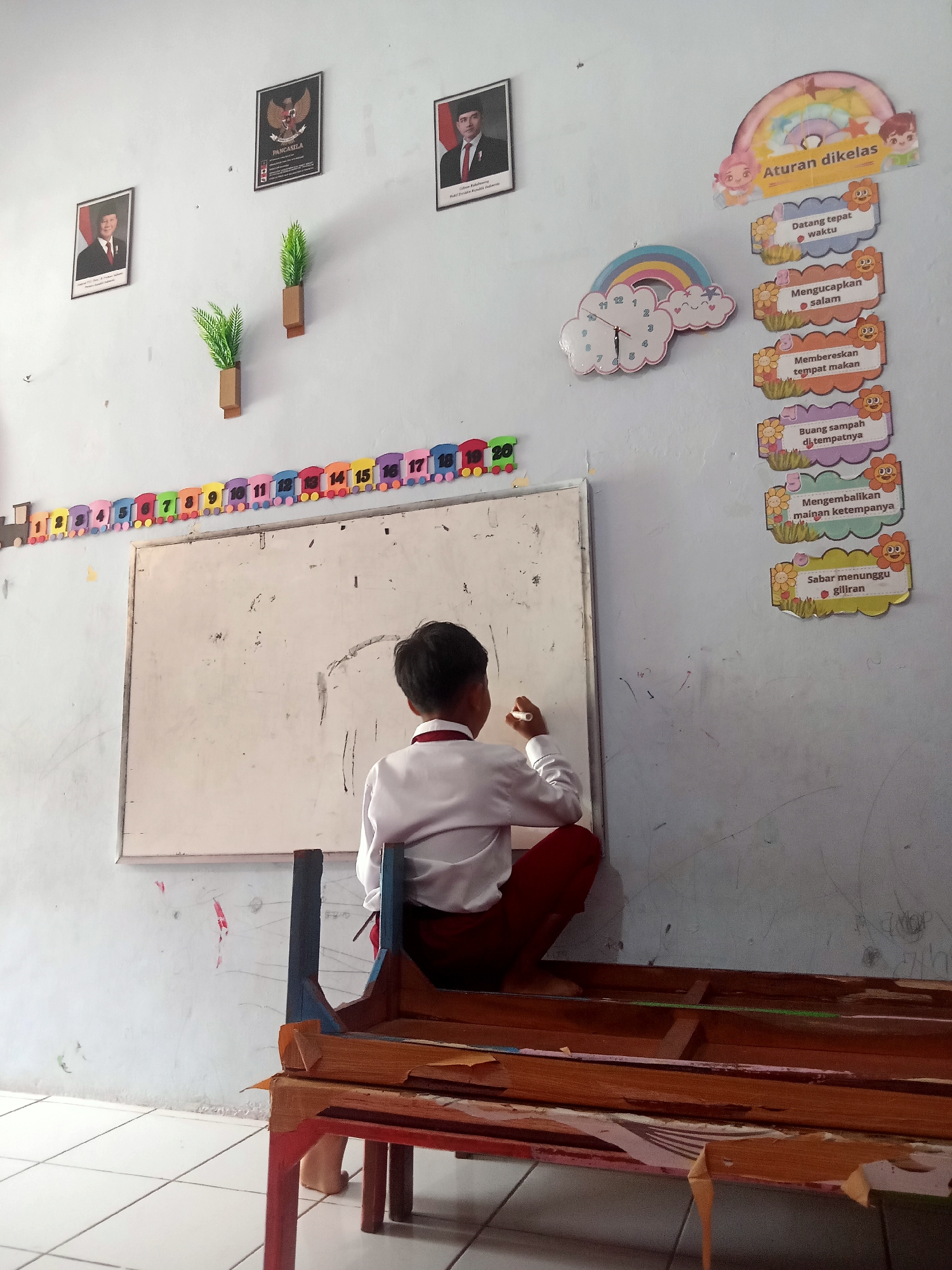 A young boy, wearing a white collared shirt and red pants, is seen from behind, drawing on a whiteboard in what appears to be a classroom. The whiteboard is mounted on a light blue wall, and a wooden bench or table with worn paint is in the foreground. Above the whiteboard, a string of colorful number cutouts shaped like train cars runs along the wall.
The wall is decorated with several items. To the left, there are two framed portraits of men, one of whom is identified as "Pancasila". Two small potted artificial plants are mounted on the wall between the portraits. To the right of the whiteboard, a decorative clock featuring a rainbow and clouds is hung, along with a series of colorful posters depicting rules or guidelines for the classroom, illustrated with cartoon children and flowers. These posters list rules such as "Datang tepat waktu" (Arrive on time), "Mengucapkan salam" (Greet), "Membereskan tempat makan" (Tidy up eating place), "Buang sampah di tempatnya" (Dispose of trash in its place), "Mengembalikan mainan ketempatnya" (Return toys to their place), and "Sabar menunggu giliran" (Patiently wait for turns).
The floor is tiled in a light color. The overall environment suggests a primary school or kindergarten classroom in a location that could be Pule, Indonesia, given the presence of Indonesian text and imagery like the Pancasila symbol. The lighting is consistent with indoor lighting.