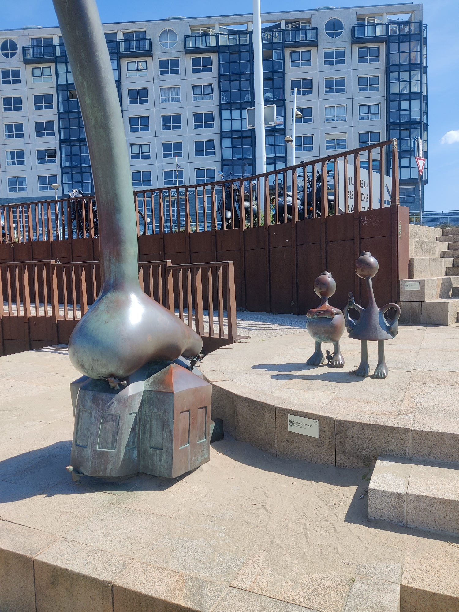 This image depicts a sculpture located at the front of the Museon museum in The Hague, Netherlands. The sculpture features a large, bronze, foot-like object resting on a building-shaped base. To the right of the main sculpture are two smaller, stylized bronze figures, resembling birds or cartoon characters. A rusted metal fence or railing runs horizontally across the middle ground. Behind the railing, a modern, multi-story building with numerous windows is visible. The scene is set outdoors on a paved area with some sand and steps leading up to the right. The sky is blue with a few white clouds, indicating a daytime setting.