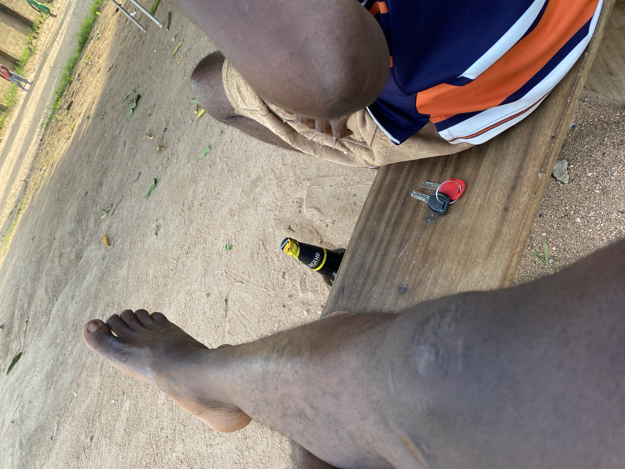 The image shows a close-up view of a person sitting outdoors, with their legs and torso prominently featured. The scene is set on a sandy ground, with scattered leaves and small pebbles visible. A wooden bench is positioned in the frame, serving as a resting place.
On the bench, two keys are placed, one with a red fob and the other a standard silver key. Next to the keys, a bottle of "LEGEND" beer is lying on its side, with its yellow cap and label clearly visible. The person is wearing a t-shirt with bold horizontal stripes in navy blue, orange, and white. Their legs are covered by light-colored shorts. The lighting suggests it is daytime, likely late afternoon due to the warm, soft light and long shadows.
The composition focuses on the immediate surroundings, with a person's legs and feet in the foreground, indicating a relaxed or casual setting. The presence of the beer bottle and keys suggests a moment of leisure or a brief stop. The overall impression is one of a simple, outdoor gathering or pause in activity. The background is blurred, but it appears to be an open area with some greenery and structures further away, possibly in Howayi, Nigeria.