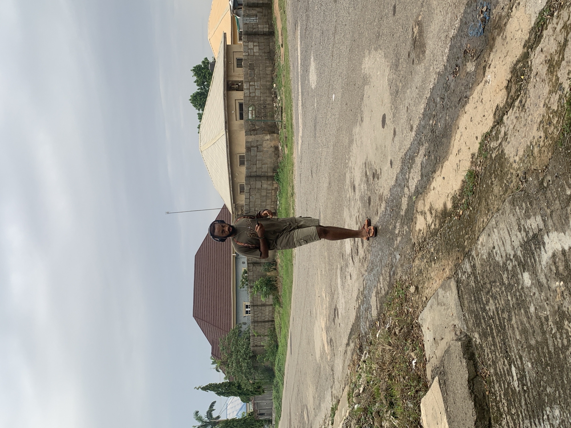 A man wearing headphones and a t-shirt is walking on a paved road in Abuja, Nigeria. He is carrying a backpack and holding a phone in his right hand. The road is bordered by a grassy verge and a concrete sidewalk. In the background, there are several houses with tiled roofs and brick walls. The sky is overcast.