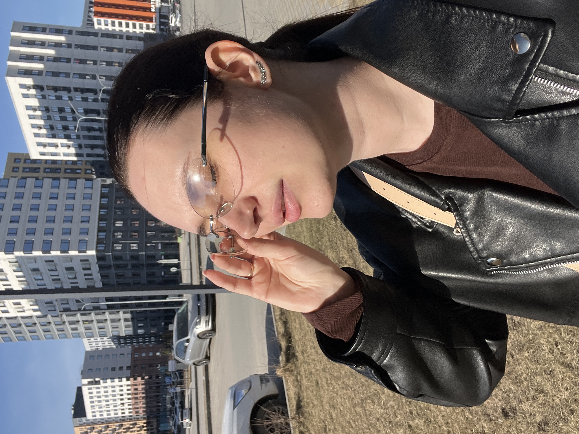 A woman with dark hair pulled back, wearing clear-framed glasses and a black leather jacket over a brown shirt, is adjusting her glasses. She is standing outdoors, likely in Tyumen, Russia, as evidenced by the presence of modern, high-rise apartment buildings with various colors and patterns in the background. There are cars parked nearby, indicating an urban or suburban street setting. The ground appears to be covered in dry grass or a similar type of ground cover. The photo seems to be taken during a sunny day.