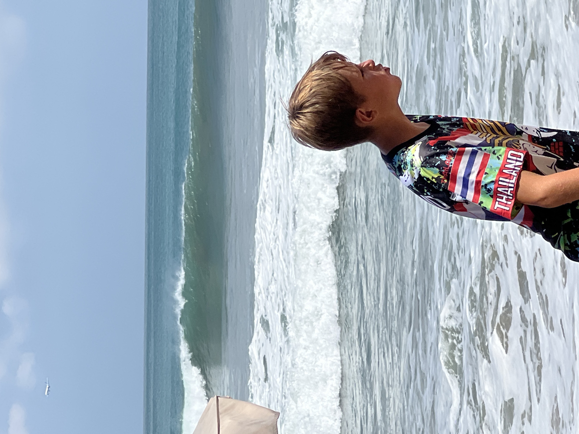 A young boy with blonde hair stands on a beach in Kuta, Indonesia. He is wearing a colorful t-shirt with the word "THAILAND" printed on it, along with other graphics. The boy is looking out towards the ocean, where waves are breaking. In the distance, an airplane is visible in the sky. A light-colored beach umbrella is partially visible on the left side of the image. The water appears to be a greenish-blue color, and the sky is a clear blue with some faint clouds.
