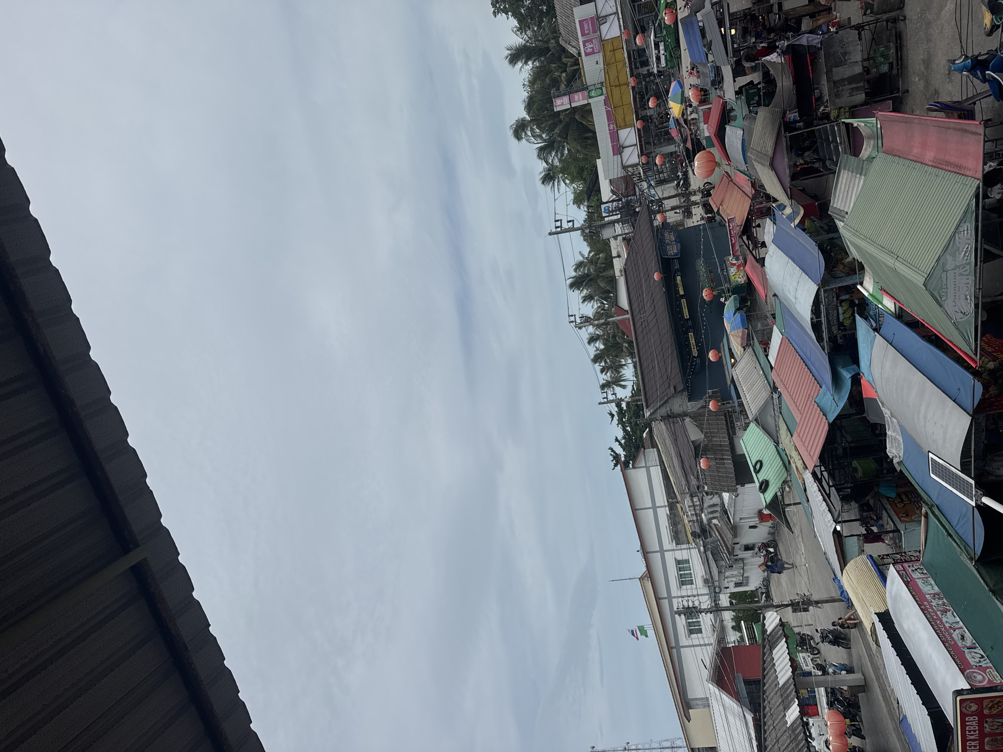 This image shows a street view in Thong Sala, Thailand. The perspective is upward, capturing the sky, buildings, trees, and market stalls. The sky is overcast. Palm trees are visible above the buildings. Market stalls with colorful awnings line the street. Some stalls have orange lanterns. Scooters are parked on the street. There appear to be people interacting among the stalls. A roof is visible in the lower left corner of the image.