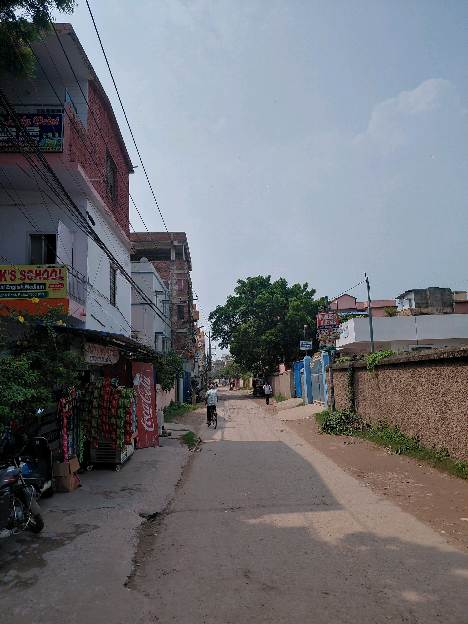 The image shows a street scene in Patna, India. On the left side of the street, there is a multi-story building with a pink and brick facade. A sign for "R.K.'S SCHOOL" with "ENGLISH MEDIUM" is visible. Below the school, there is a small shop with various products displayed outside, including bags of snacks and a Coca-Cola advertisement. A scooter is parked in front of the shop. Further down the street on the left, there are more buildings and a person walking.
On the right side of the street, there is a textured brown wall. Beyond the wall, there are more buildings, including one with a light pink facade and another with a white exterior. A person on a bicycle is visible in the middle of the street, cycling away from the viewer. A rickshaw is also present further down the street on the left. The sky is mostly clear with some clouds. The road is paved and appears to be in moderate condition with some cracks and unevenness. Trees are visible on the right side of the street and in the background.