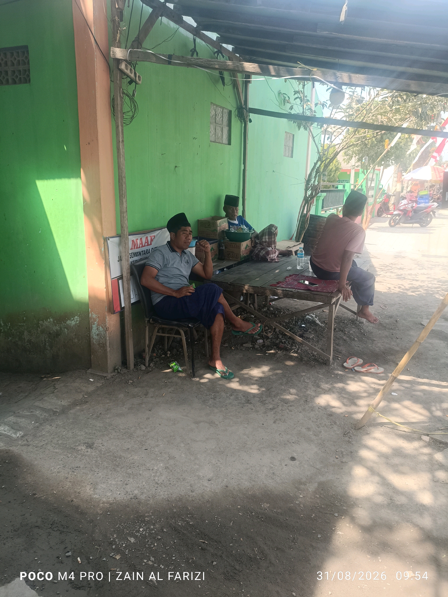The image depicts an outdoor scene in Cendagah, Indonesia, under a wooden-framed roof casting shadows. In the foreground, a man wearing a grey polo shirt, dark blue pants, and a black peci (cap) sits on a metal chair, holding a cup and appearing to be in conversation. To his right, another man in a maroon shirt and dark pants sits on a wooden table. Further back, another individual in a blue shirt and peci is seated. A table is laden with various items, including a water bottle, a red mat, and what appear to be boxes. The background shows a green building with barred windows, trees, a street with a motorbike, and a market stall. The ground is paved with concrete and dirt. The timestamp "31/08/2026 09:54" is visible in the lower right corner.