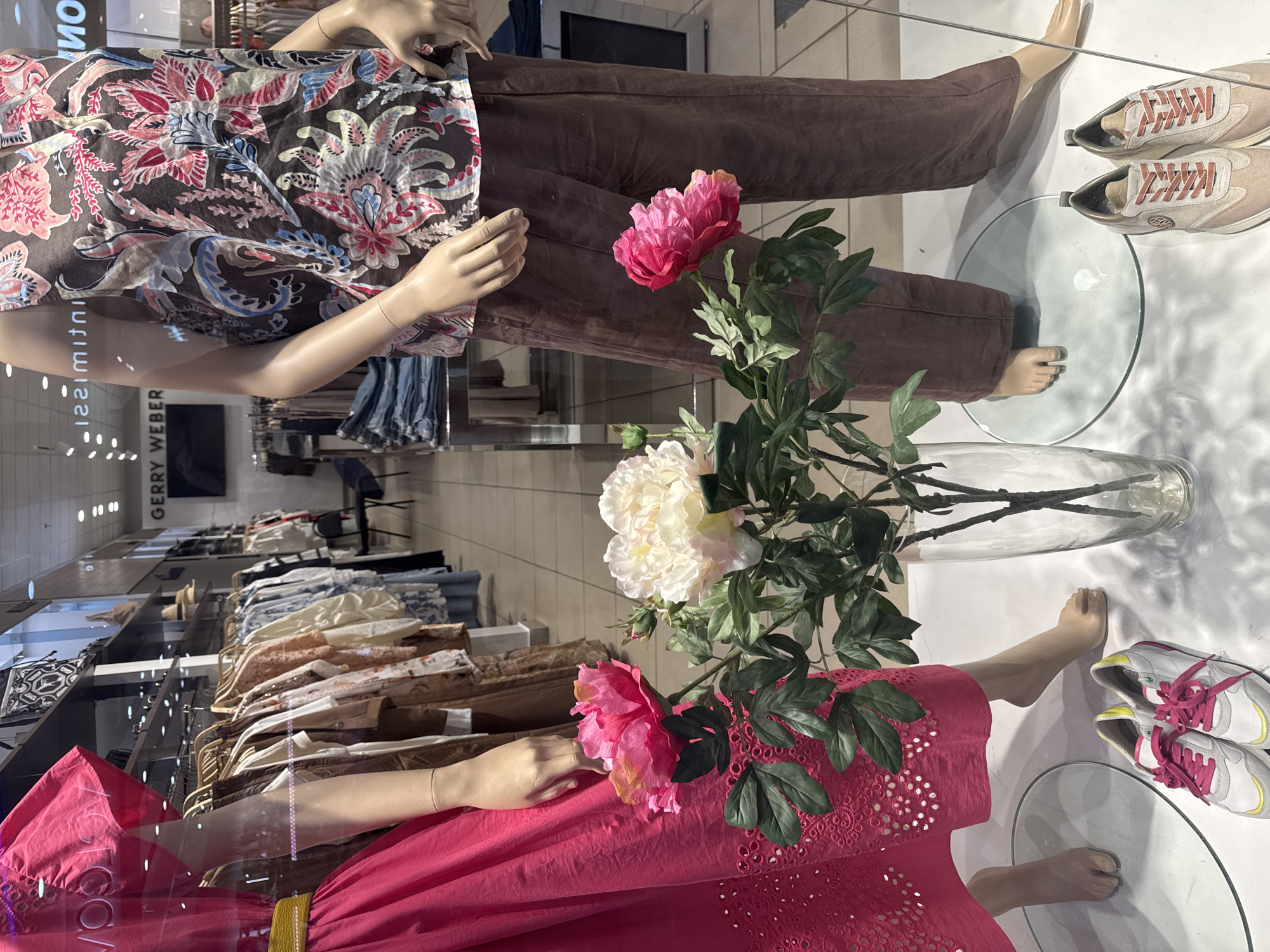 The image shows a store window display. Two mannequins are visible, each wearing different outfits. The mannequin on the top wears a brown patterned shirt and brown pants, paired with beige sneakers. The mannequin below is dressed in a bright pink dress. Between them is a clear vase holding flowers. The display is on a white surface. Outside the window, clothes on hangers are seen in the shop interior.